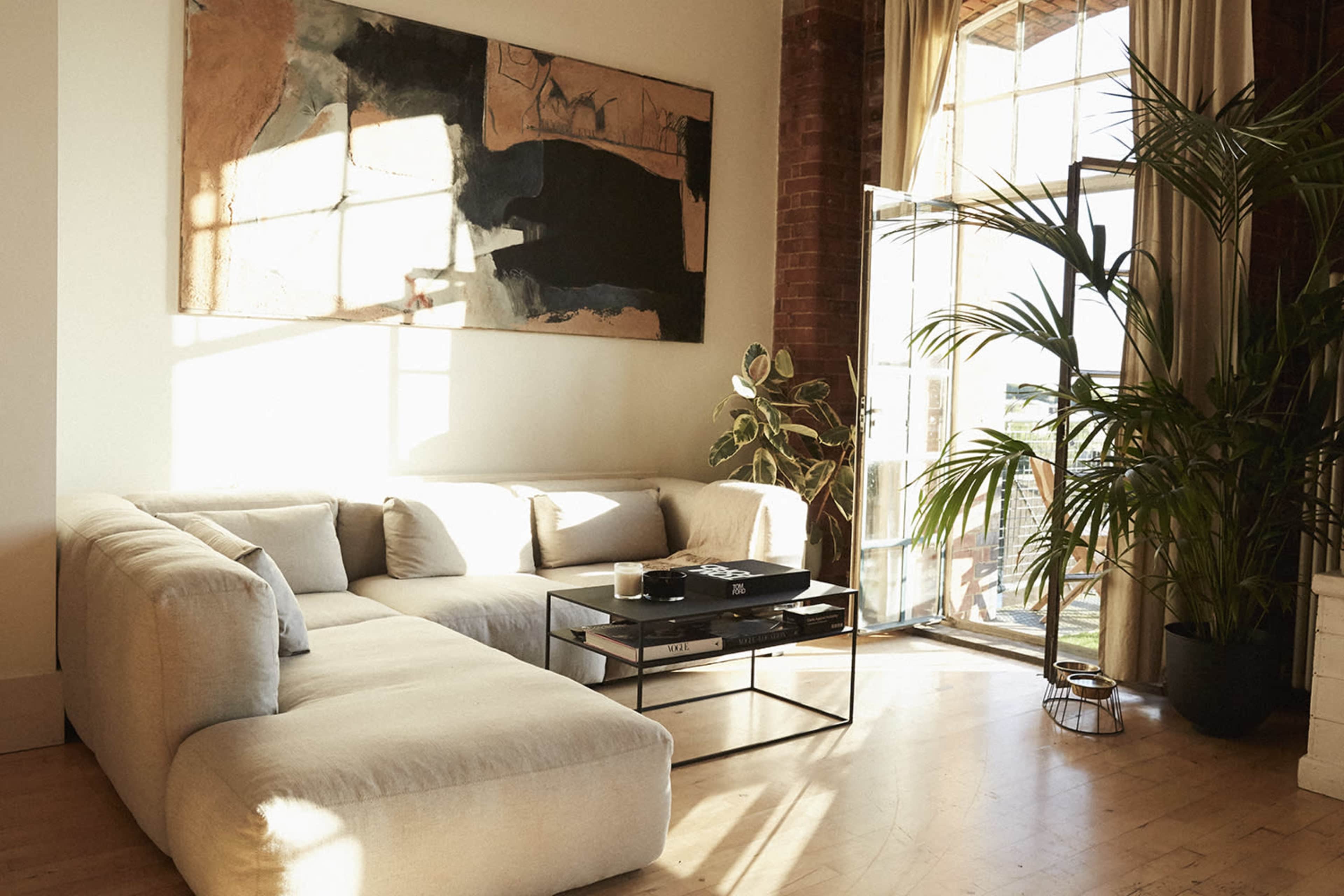 A bright living room features a large sectional sofa, a coffee table with decor items, and a tall plant next to wide windows opening to a balcony.
