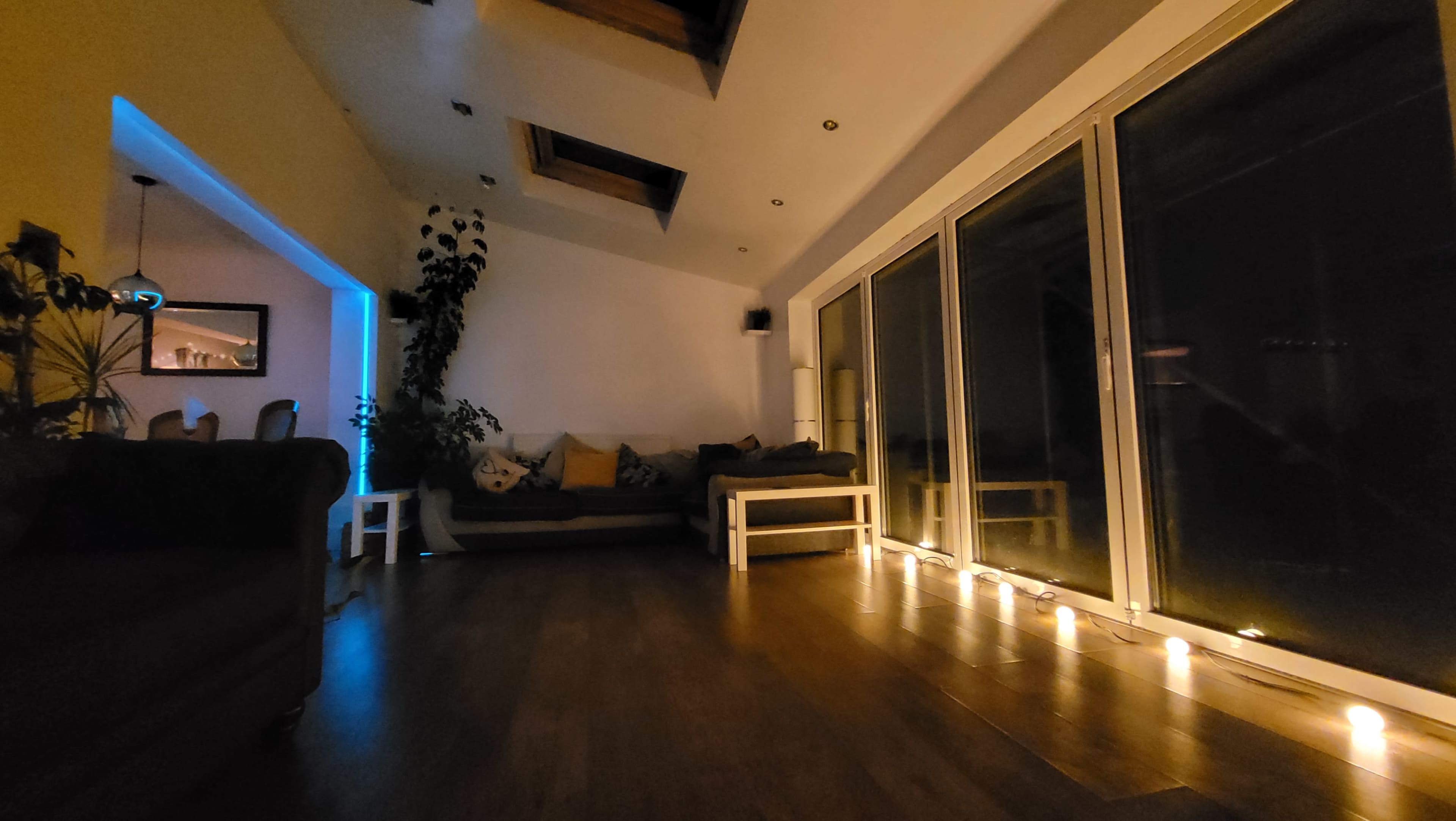 A spacious interior features a seating area with a sofa, illuminated by candles along the floor and soft blue light from a nearby room.