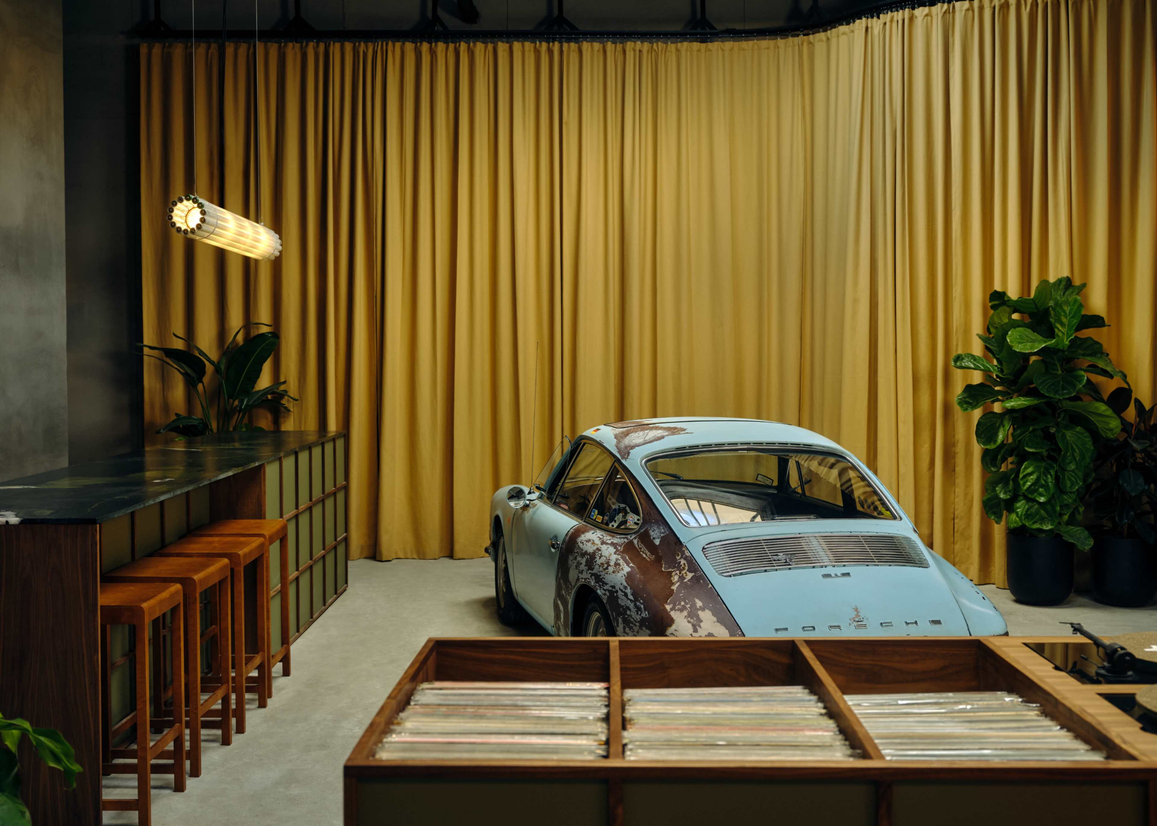A vintage blue Porsche is positioned next to a bar with wooden stools, set against a backdrop of yellow curtains and greenery.