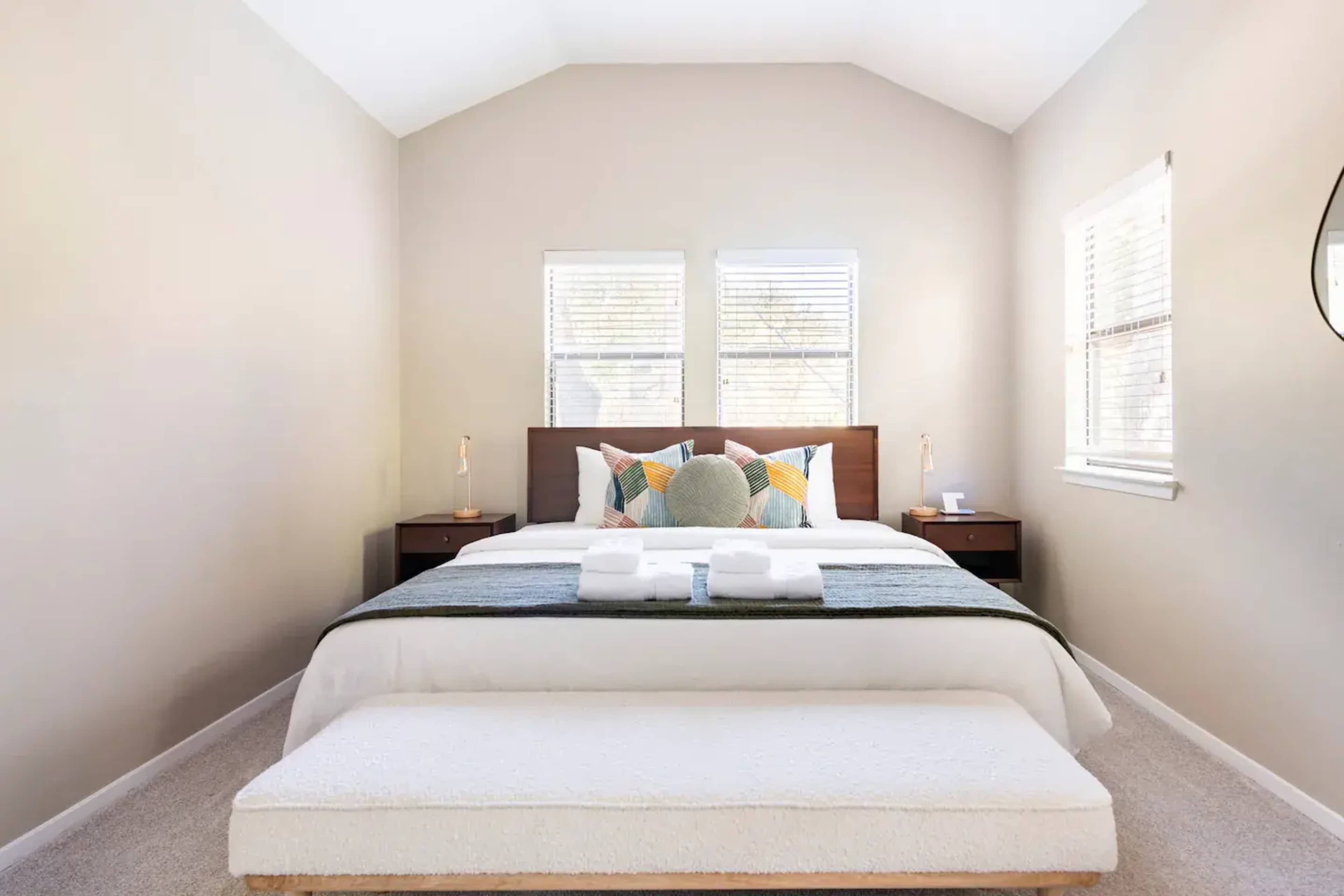 The image shows a neatly arranged bedroom with a large bed, two side tables, and large windows allowing natural light.