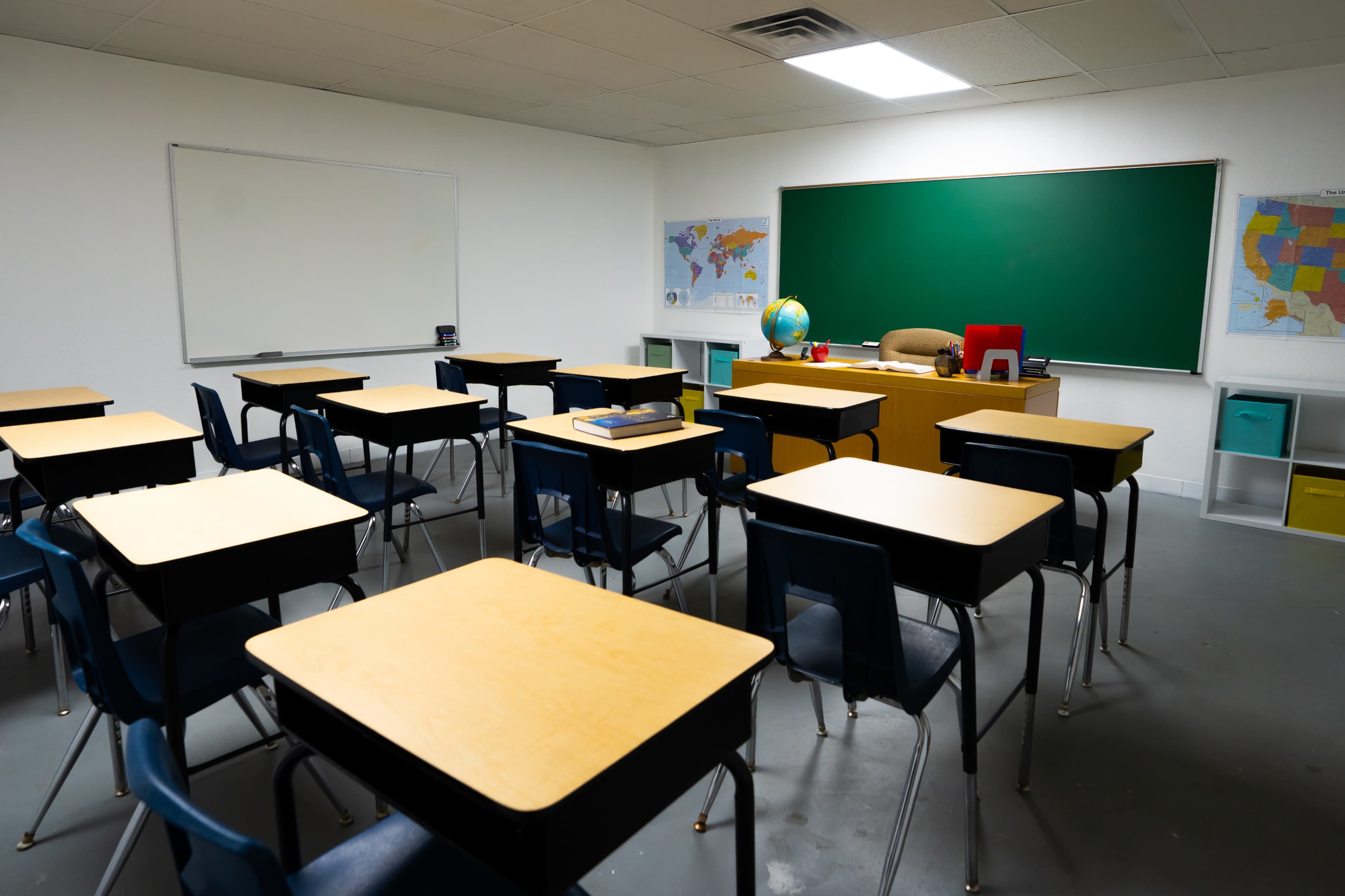 A classroom is arranged with empty wooden desks facing a green chalkboard and a teacher's desk at the front.