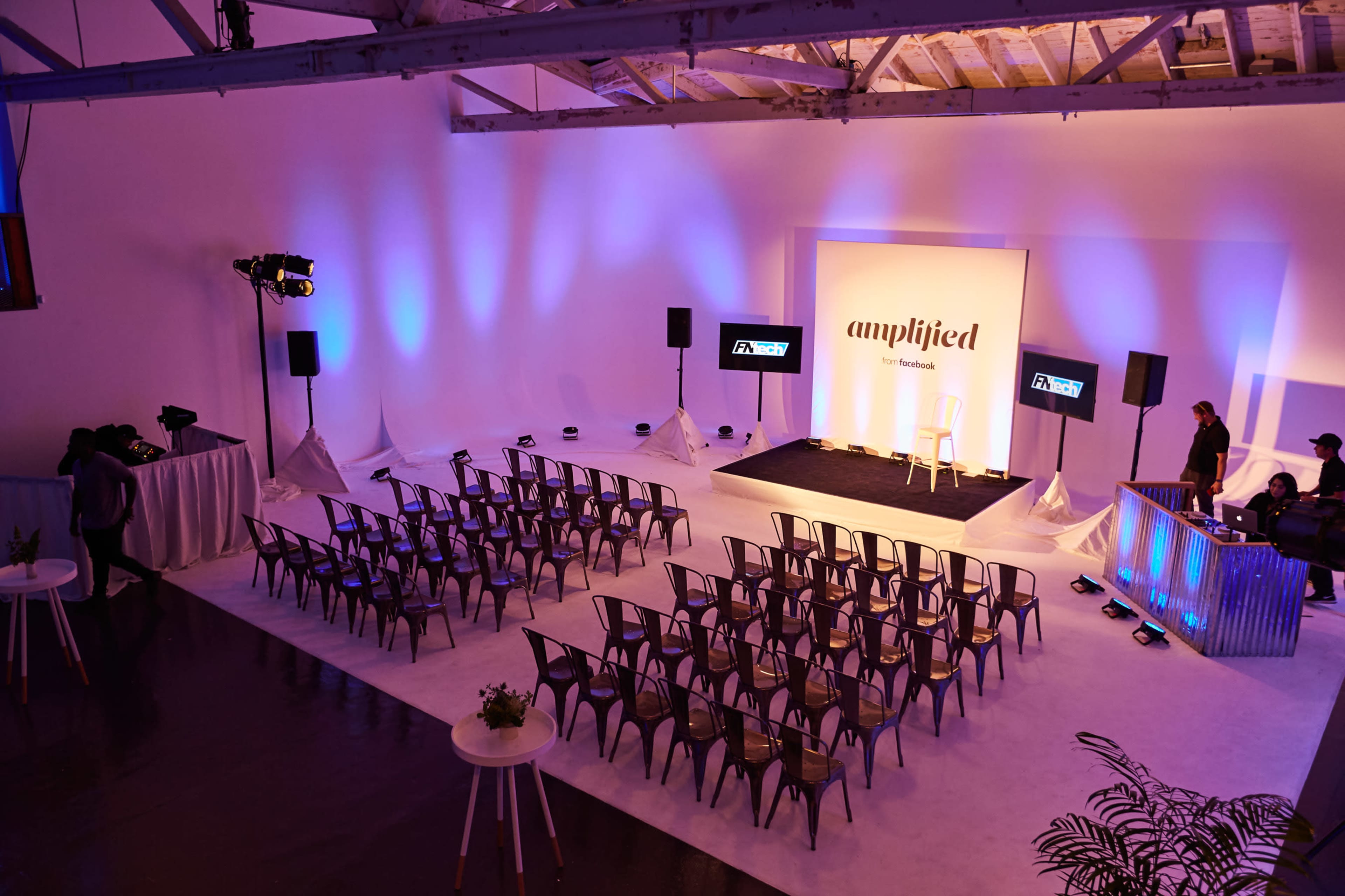 The image shows an event space set up with rows of metal chairs facing a stage, adorned with purple lighting and a backdrop that reads "amplified."