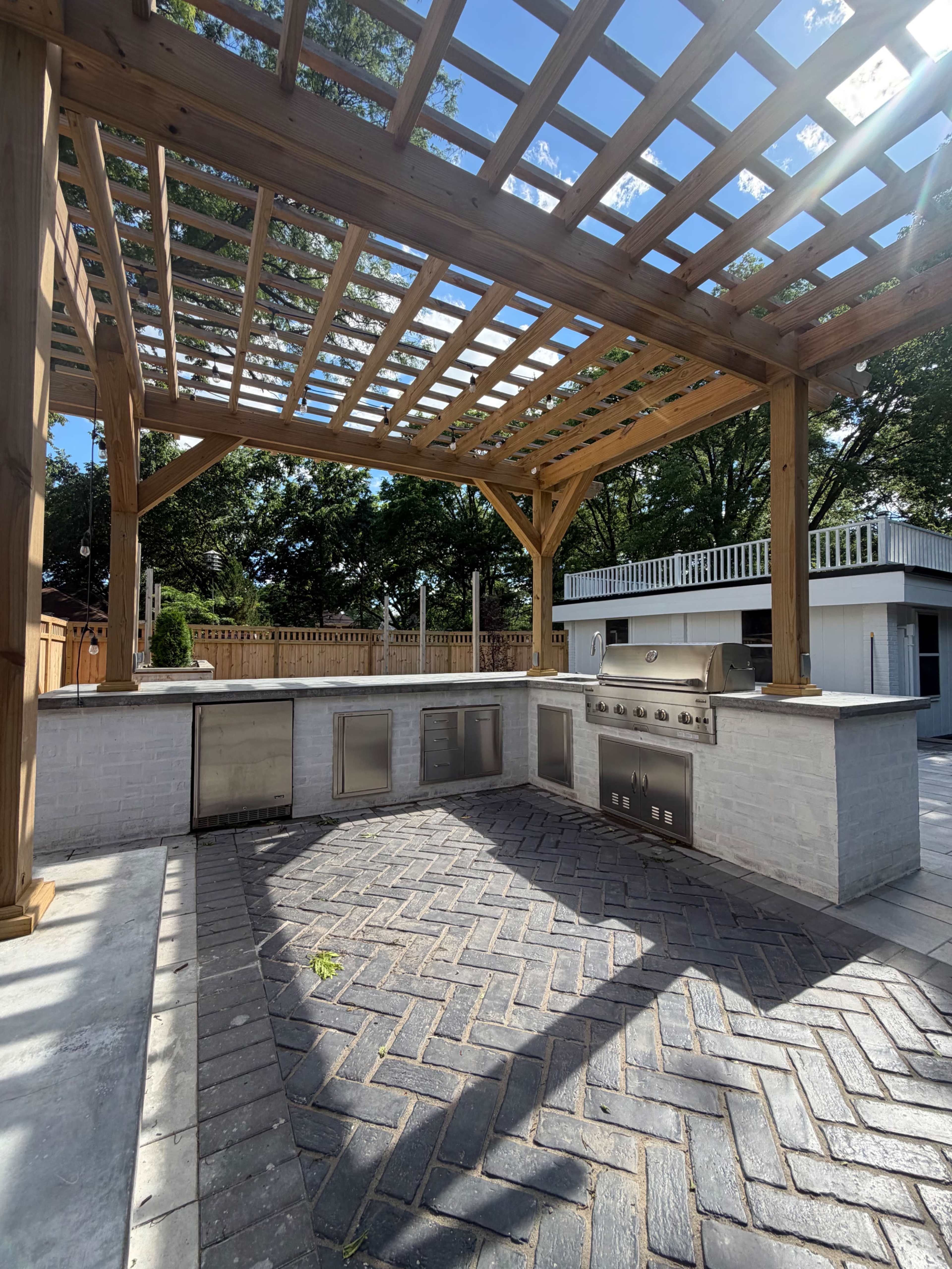 The image shows an outdoor kitchen area featuring a stone counter with built-in appliances, surrounded by a wooden pergola and a patterned stone patio.