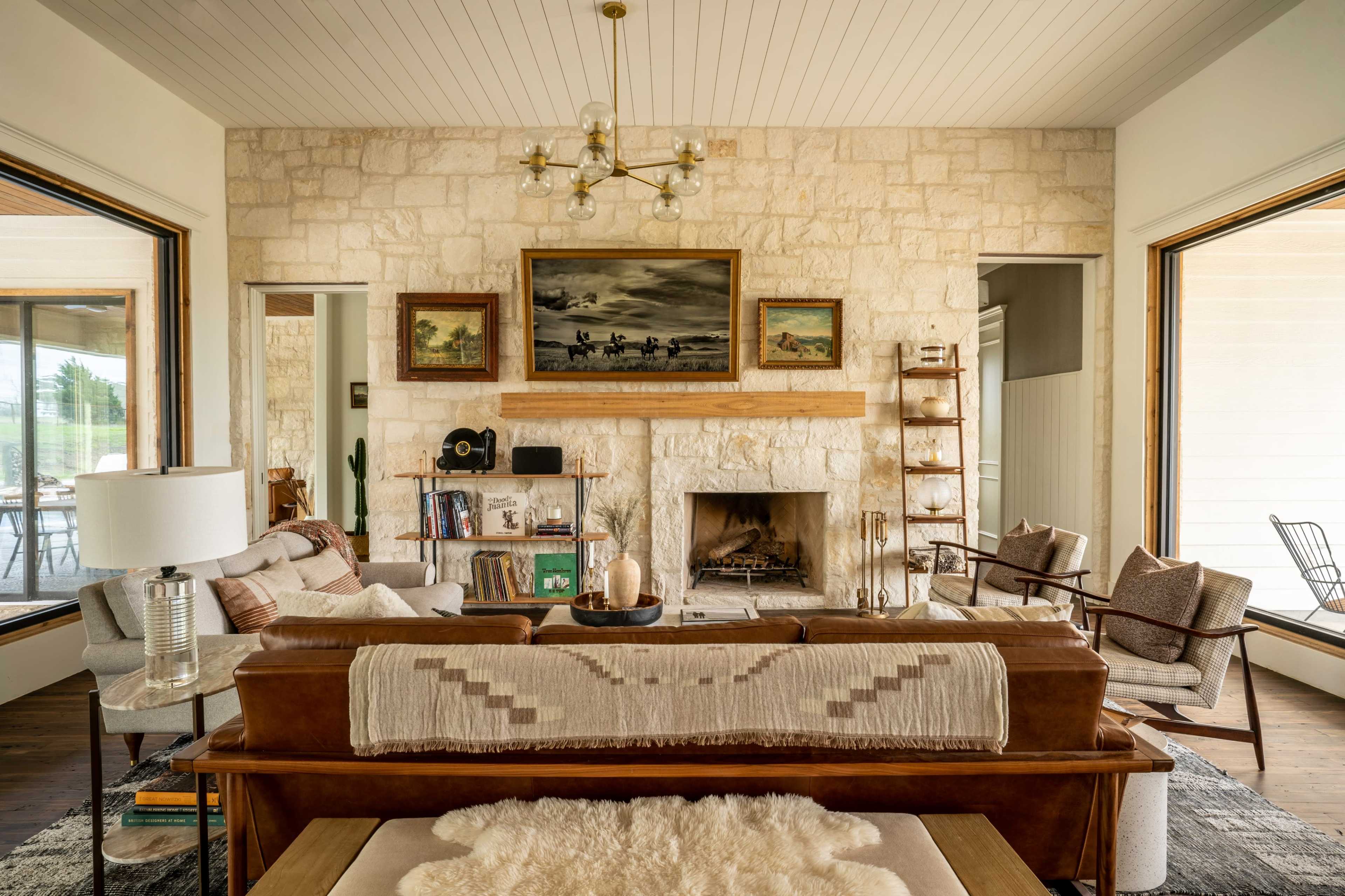 The image shows a modern living room featuring a stone fireplace, large windows, a combination of wood and upholstered furniture, and mounted artwork on the walls.