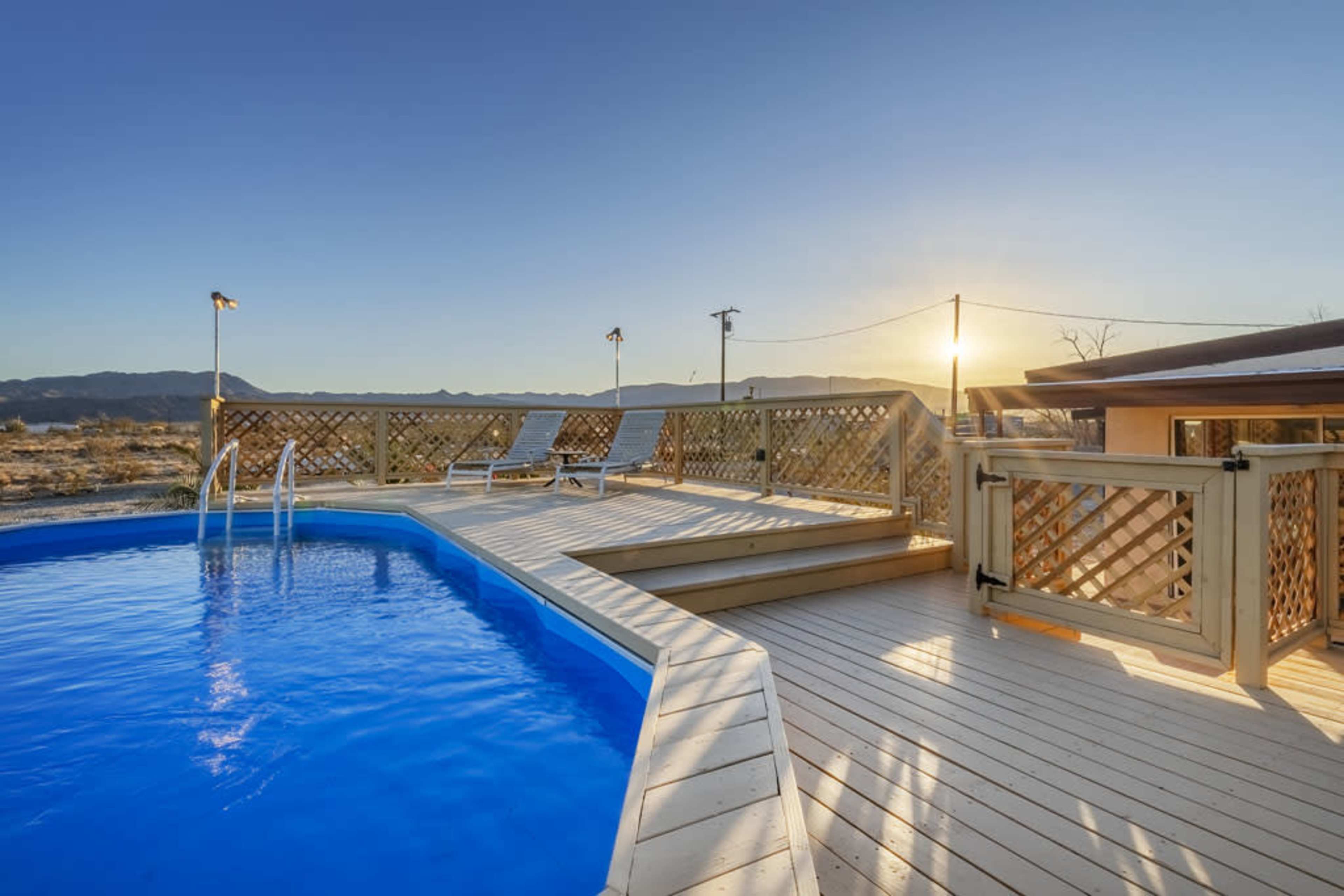 A clear blue pool is surrounded by a wooden deck, with a lounge chair positioned nearby, as the sun sets over distant mountains.