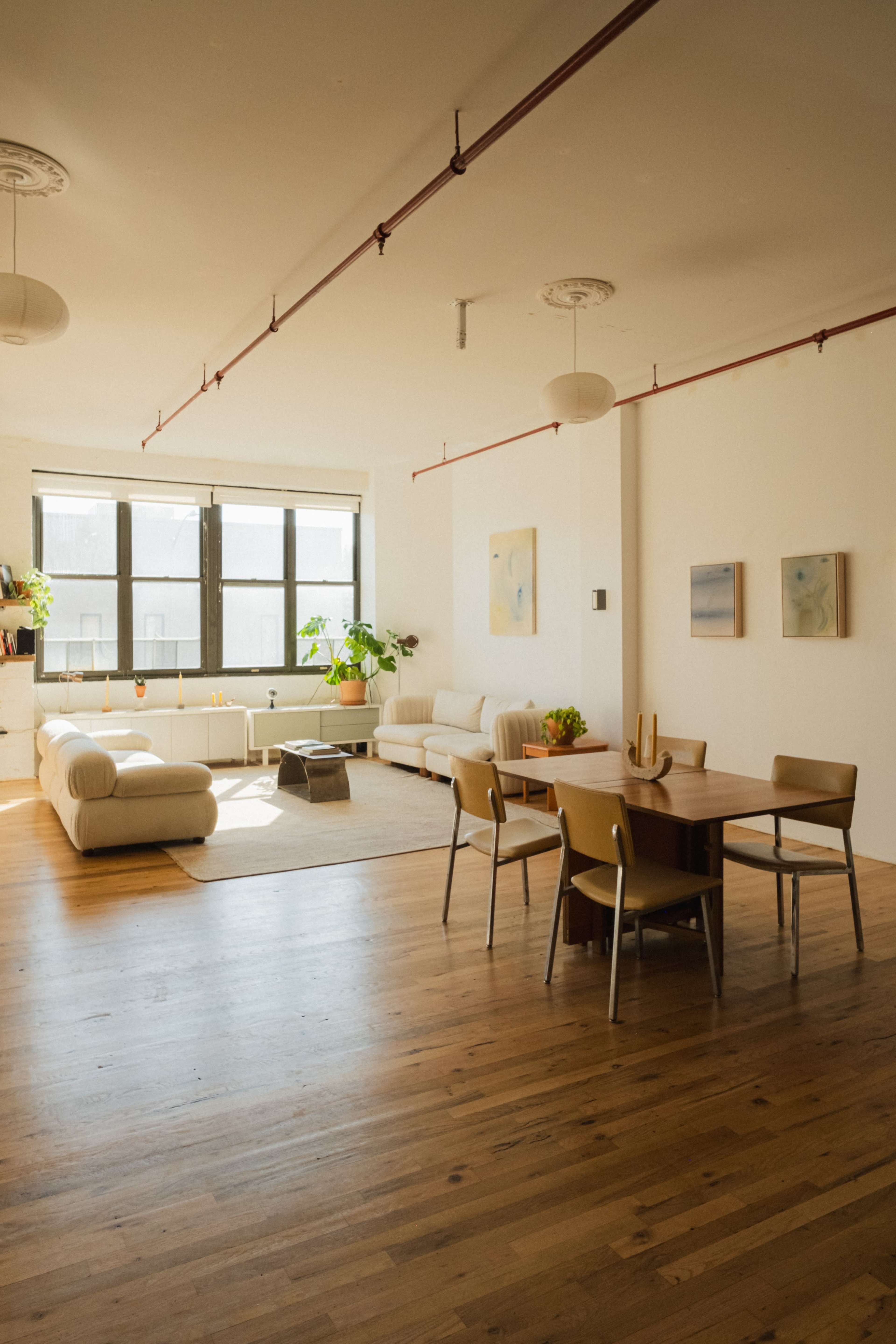 The image shows a minimalistic living space with wooden flooring, two sofas, a dining table with chairs, and large windows allowing natural light to fill the room.