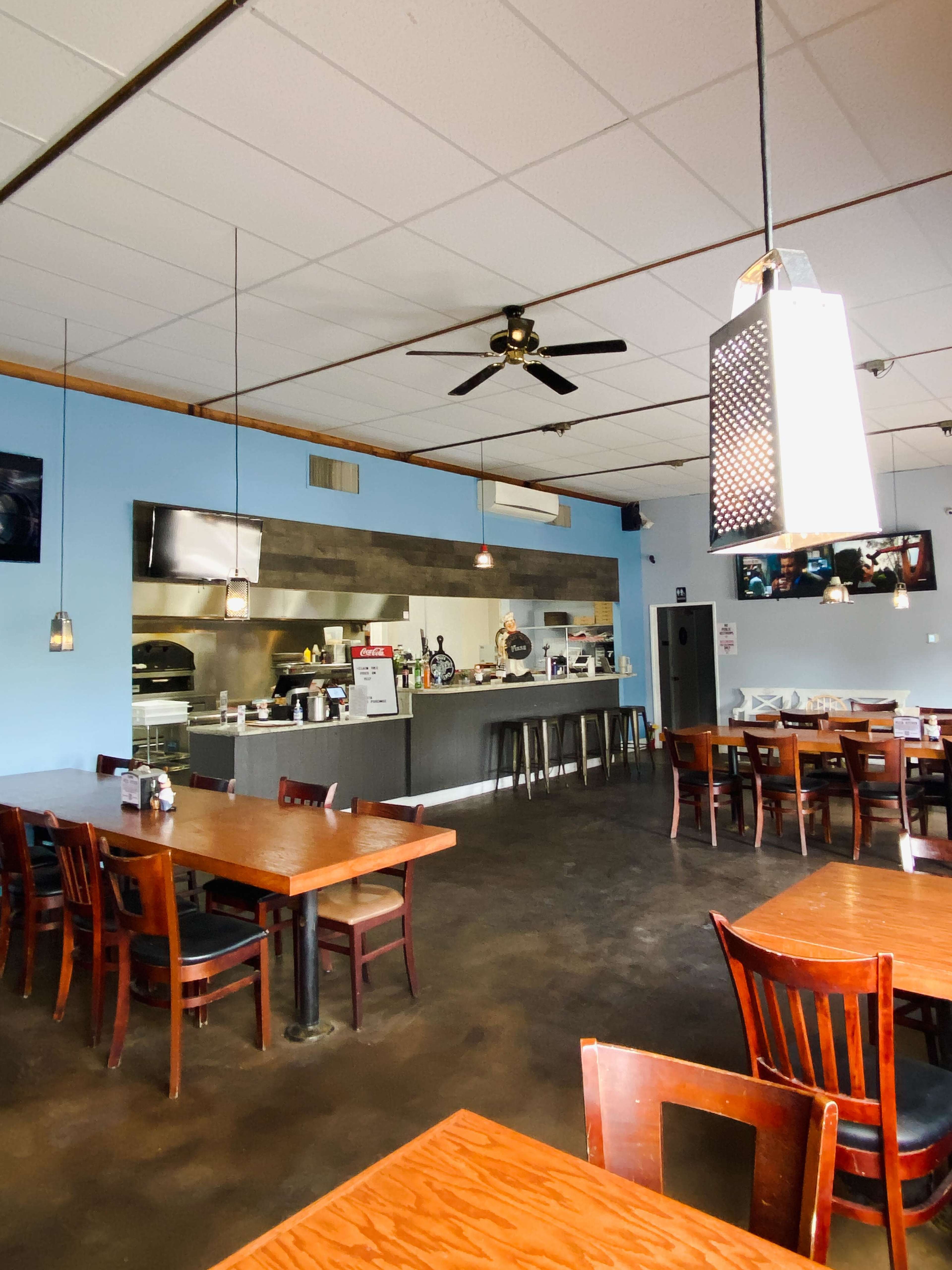 A restaurant interior with wooden tables and chairs, a counter in the back, and a kitchen area behind a stainless steel panel.