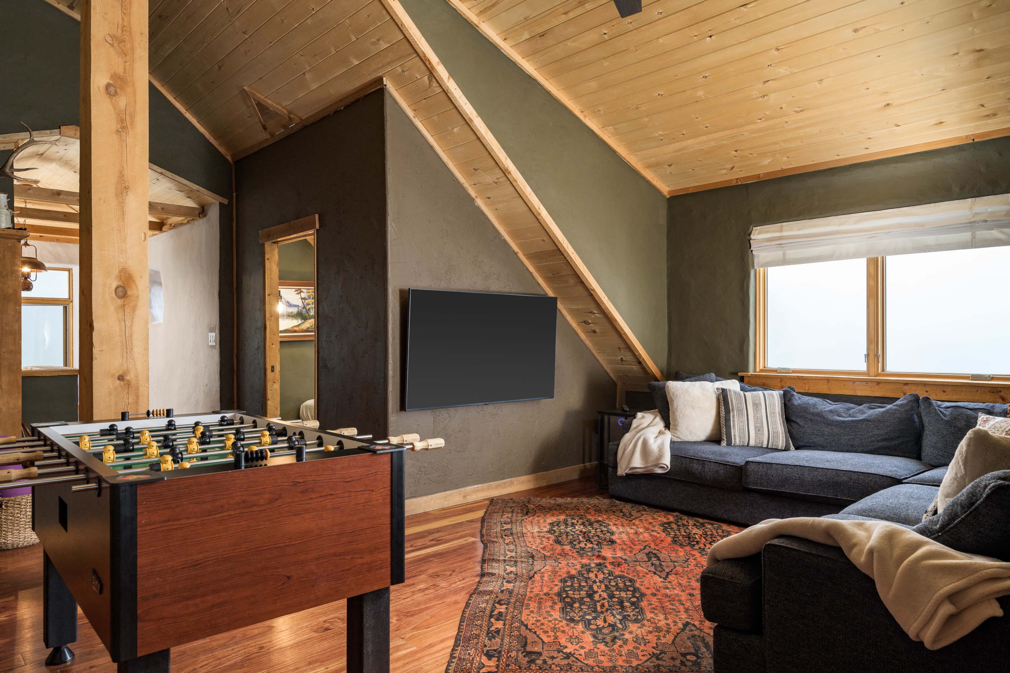 A cozy living space features a foosball table, sofa, and a large window with views, complemented by a wooden ceiling and earthy wall colors.