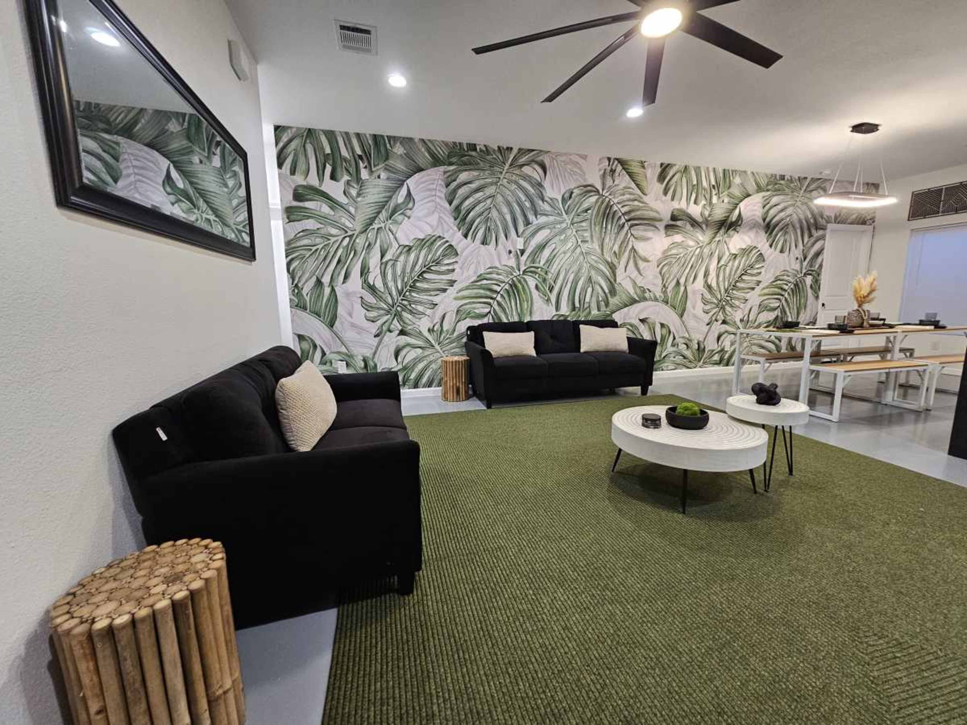 The scene features a modern living room with black sofas, a round coffee table, a green area rug, and a large tropical leaf mural on the wall.