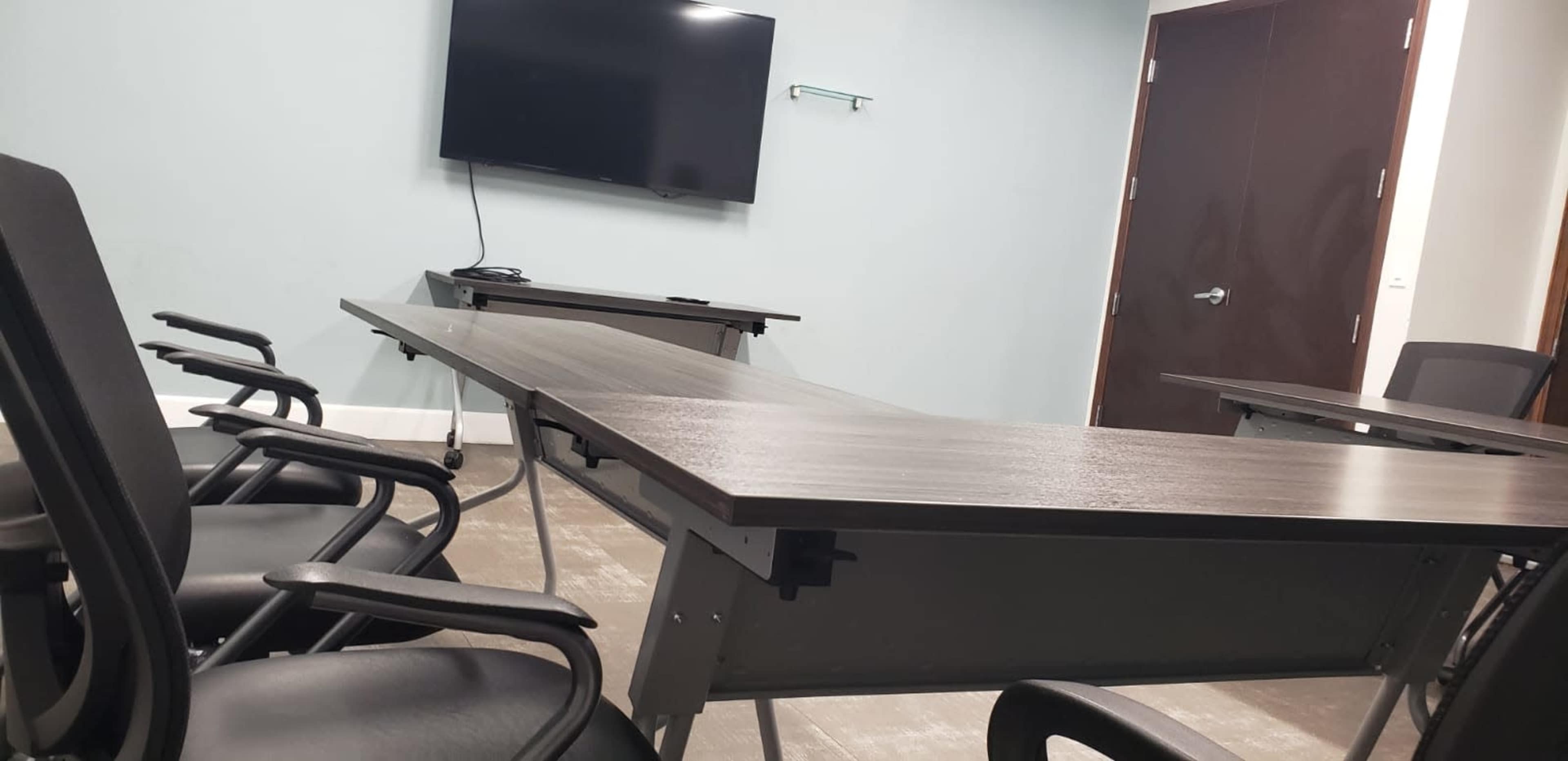 A conference room with a large table and several black office chairs, featuring a wall-mounted TV and a door.