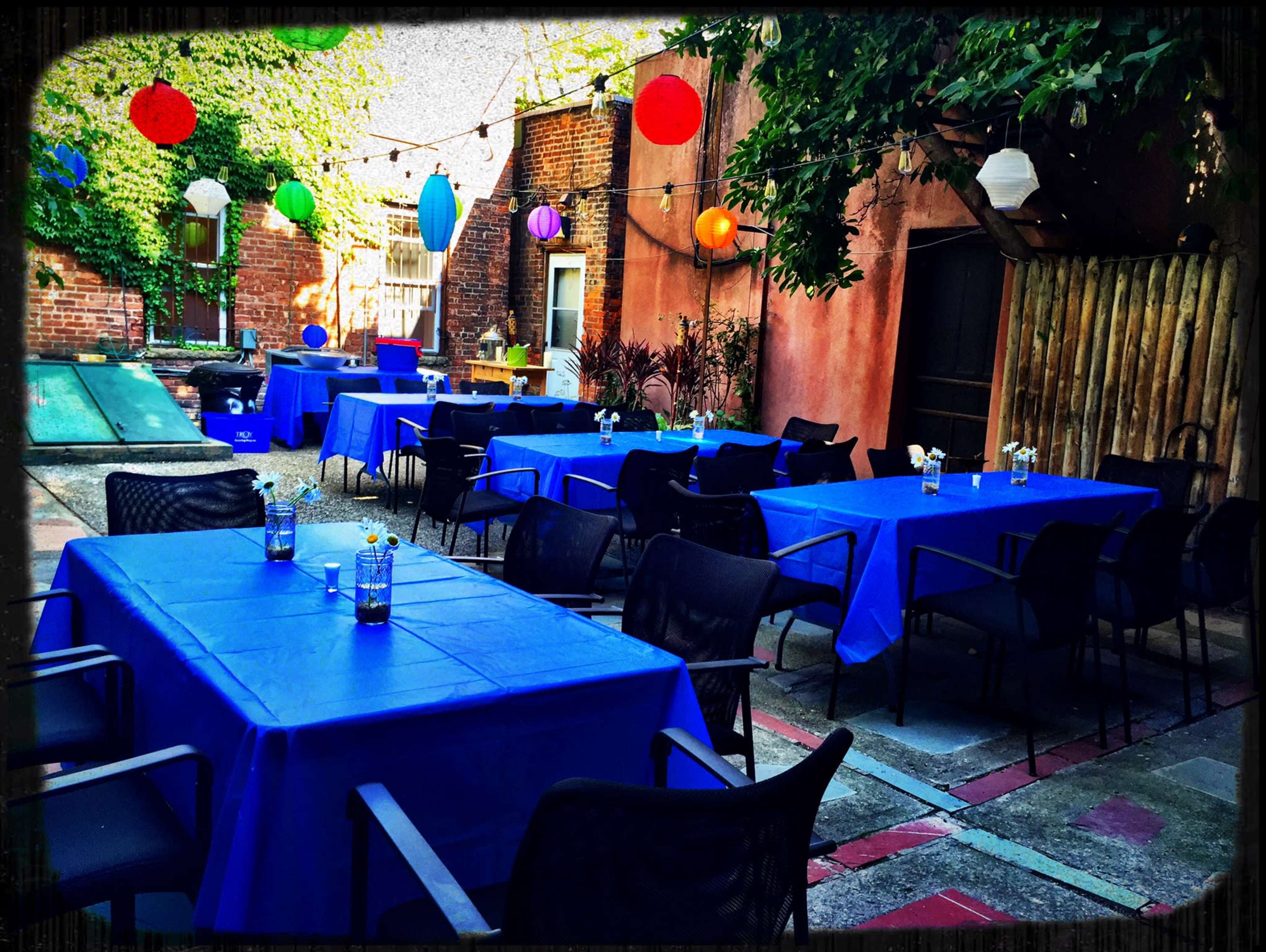 An outdoor area features several blue-covered tables arranged neatly, surrounded by colorful lanterns and string lights overhead.