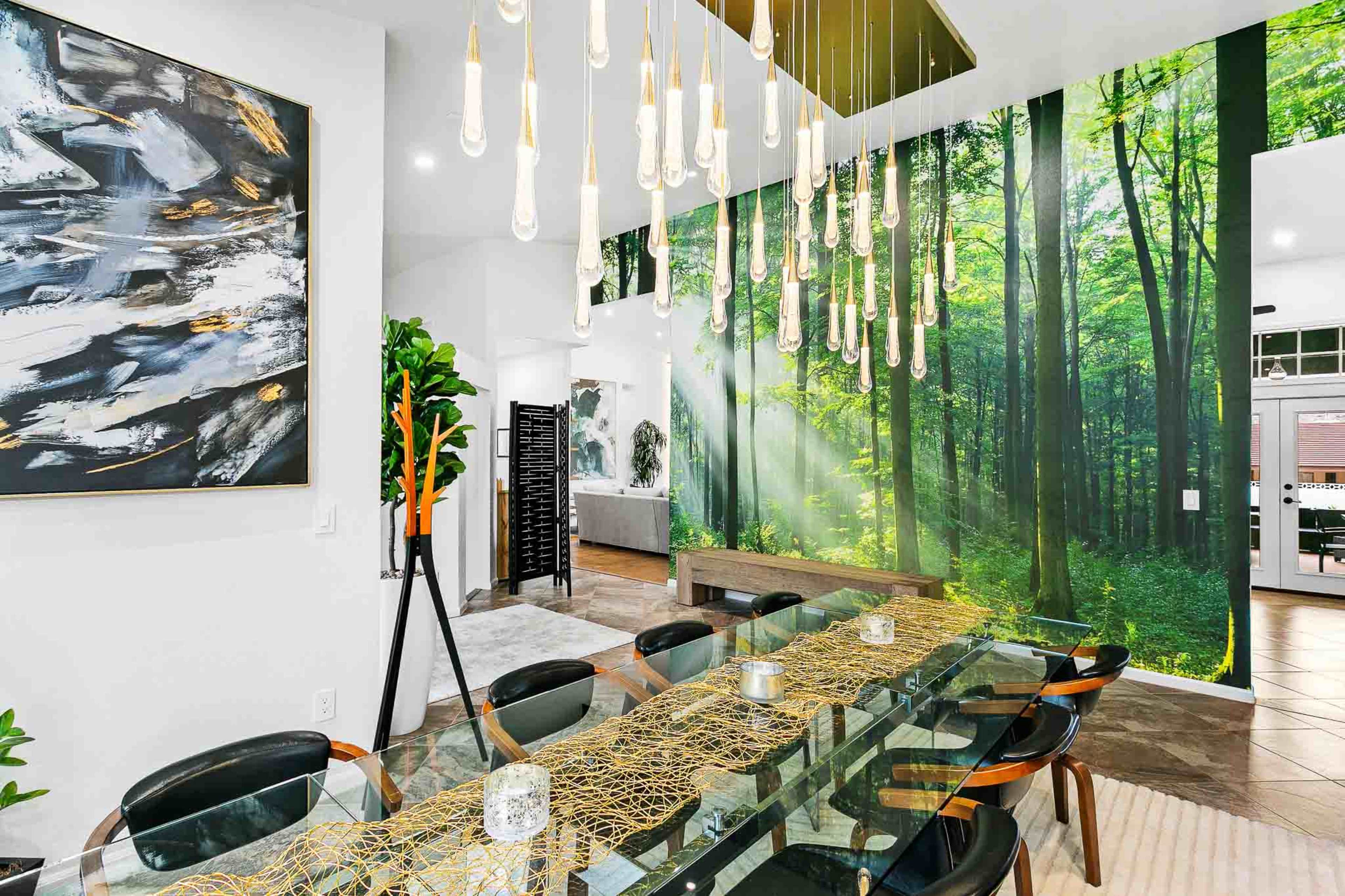 A modern dining area features a long glass table set with decorative elements, surrounded by black chairs, with a forest-themed mural on one wall and unique pendant lighting above.