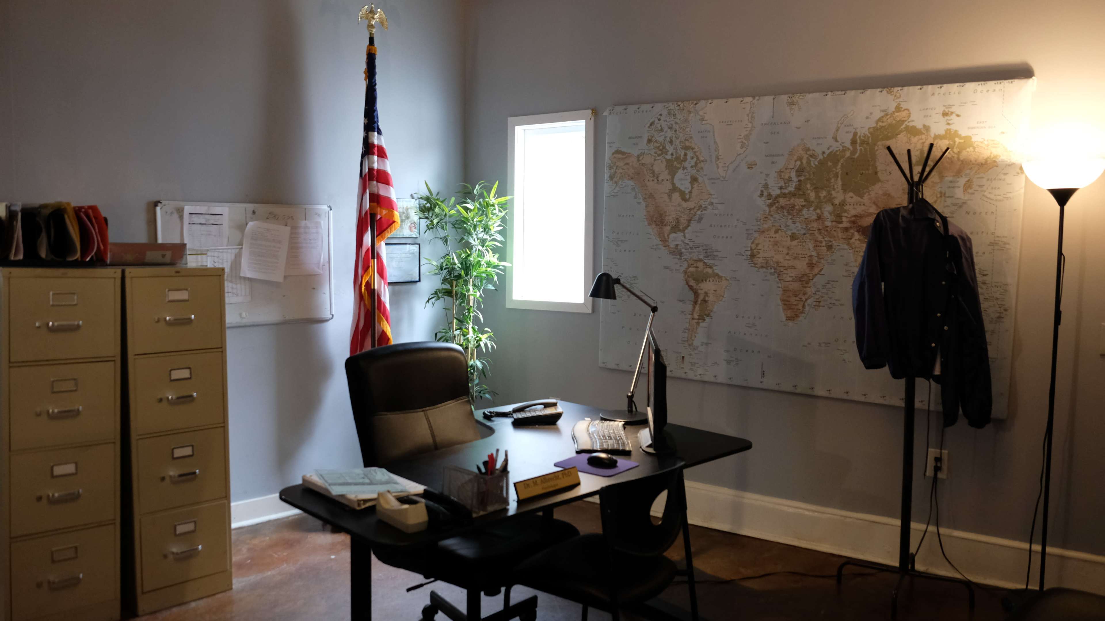 The image shows an office space with a desk, an American flag, a world map on the wall, and a plant beside a window.