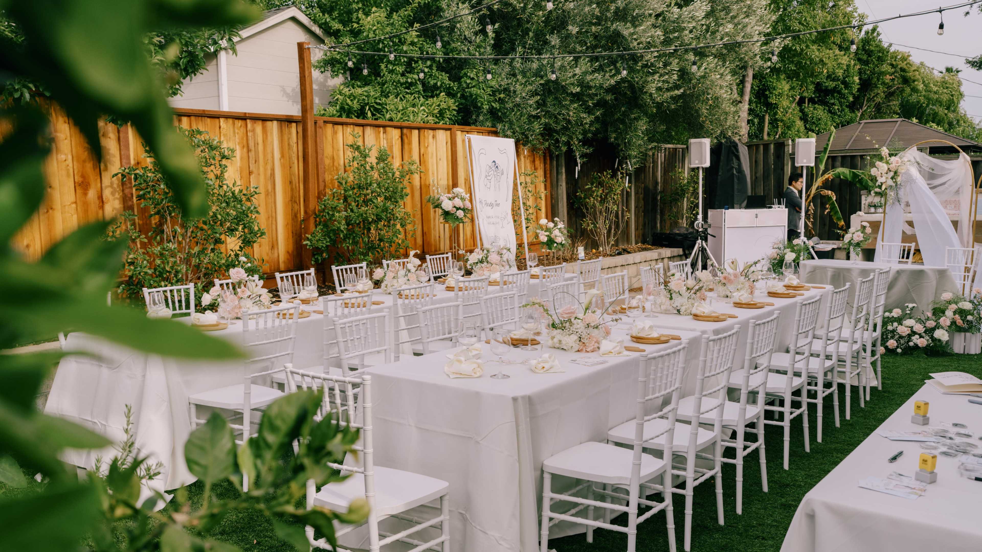 A neatly arranged outdoor wedding reception features long tables draped in white tablecloths set with centerpieces and surrounded by chairs, against a backdrop of greenery and wooden fencing.