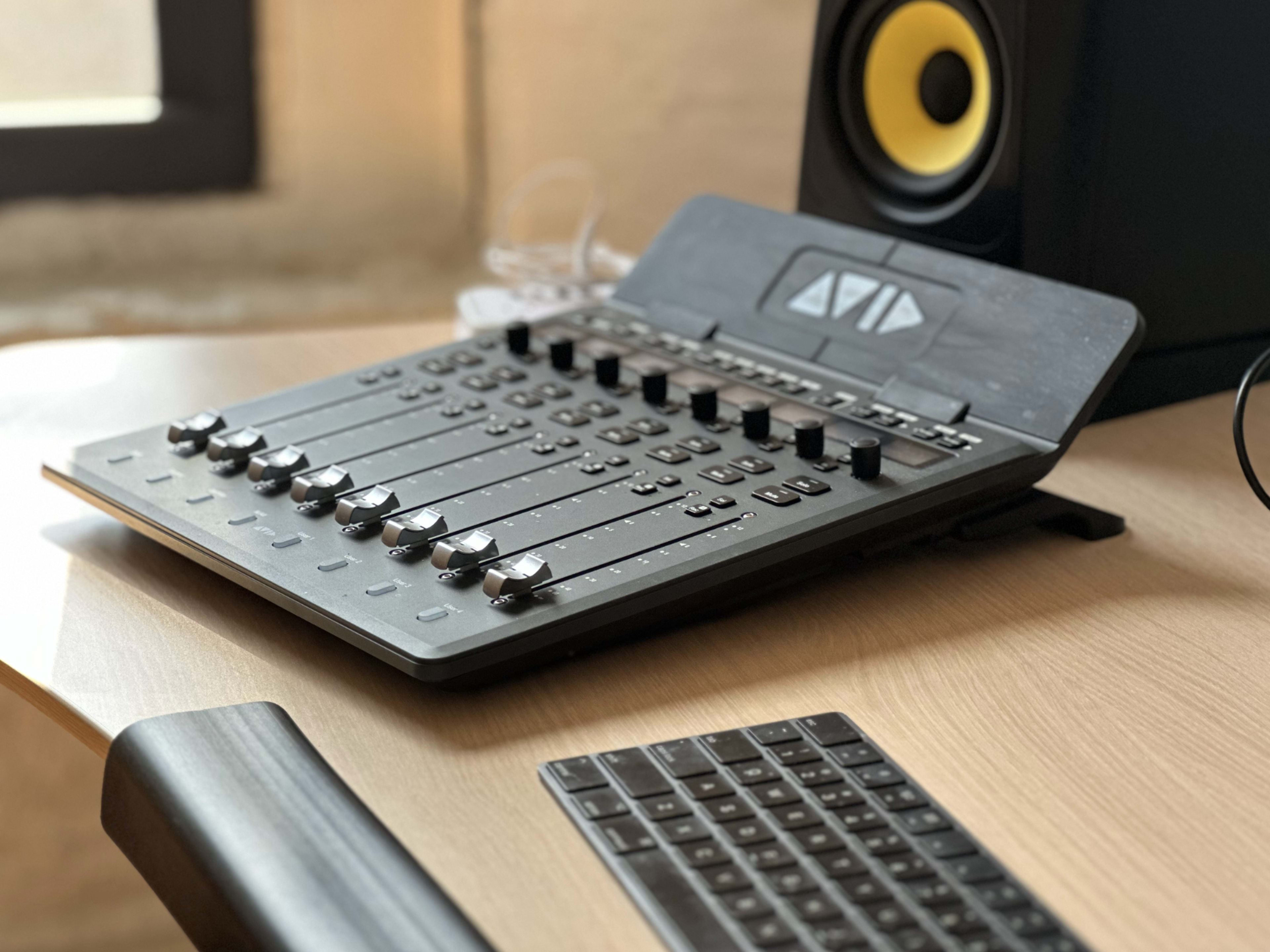 A digital audio mixer with multiple sliders and knobs is placed on a wooden desk next to a computer keyboard, with a studio monitor in the background.