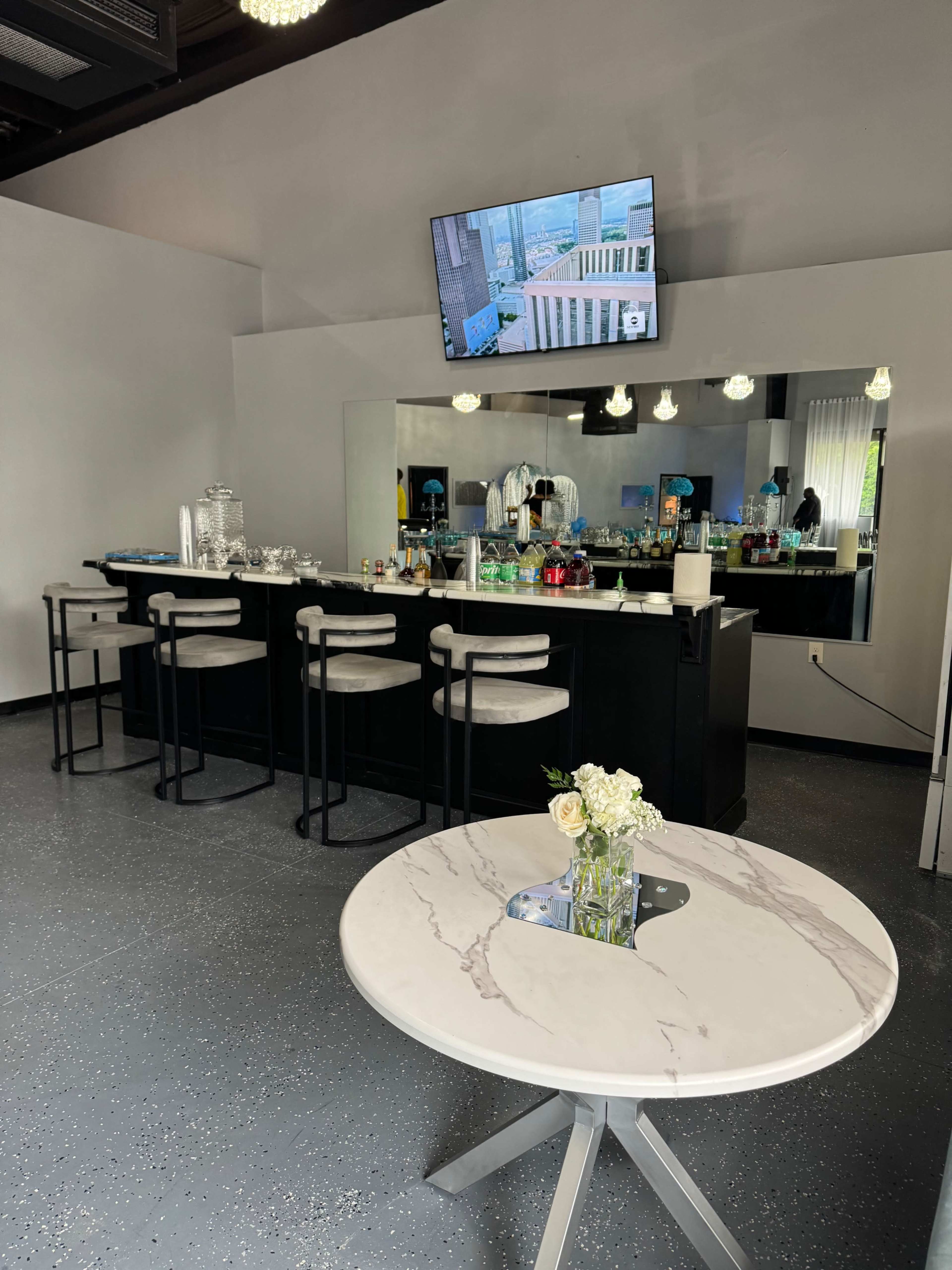 A modern bar area features a countertop with stools, a mirror behind the bar, and a television mounted on the wall, alongside a small table with a vase of flowers.