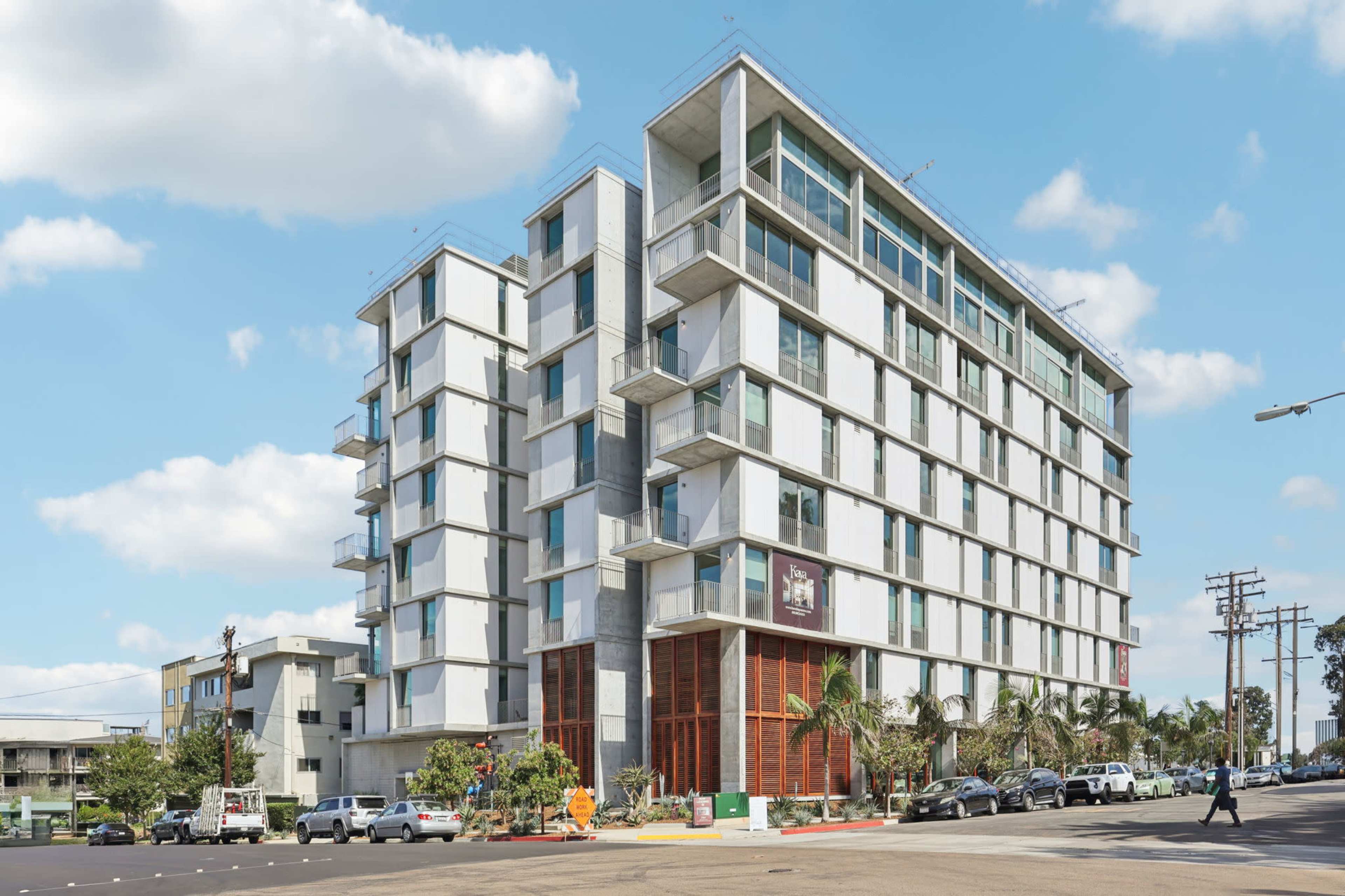 A modern, multi-story building with a glass facade and balconies stands at the corner of a street, surrounded by trees and parked vehicles.