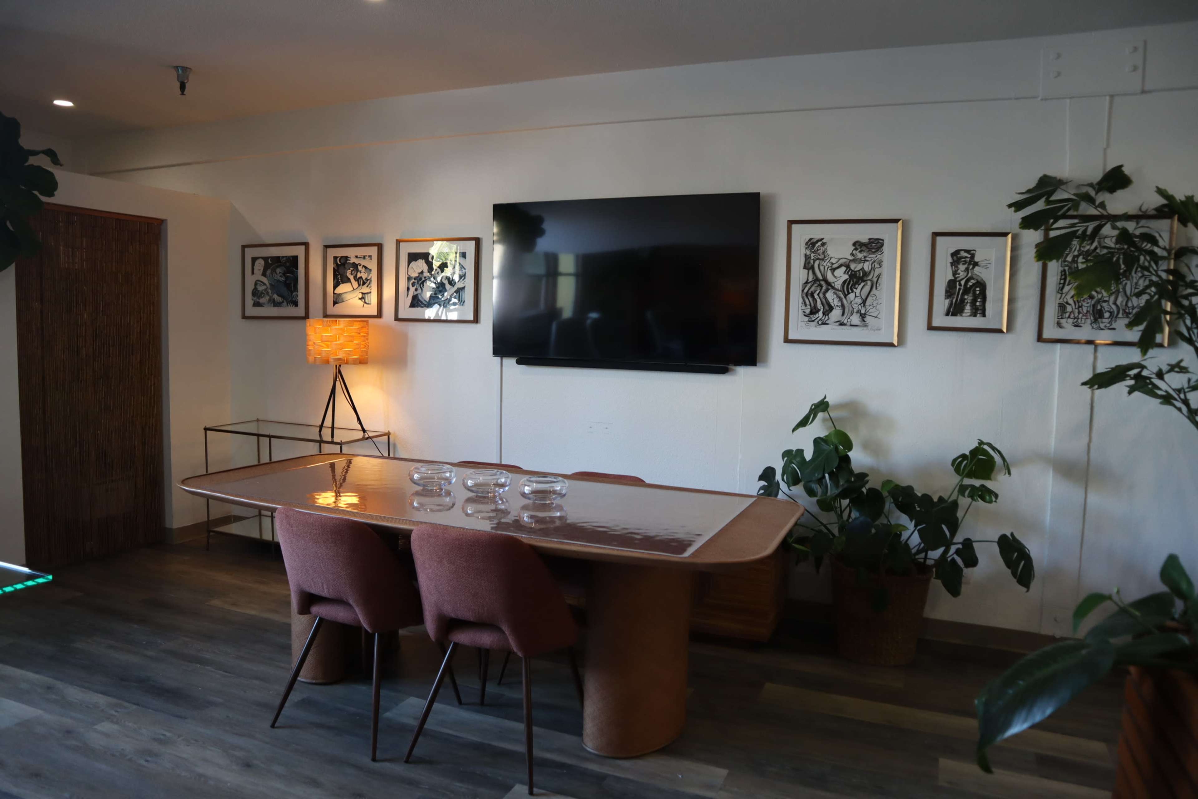 A modern conference room features a wooden table with pink chairs, surrounded by framed artwork on the walls and indoor plants.