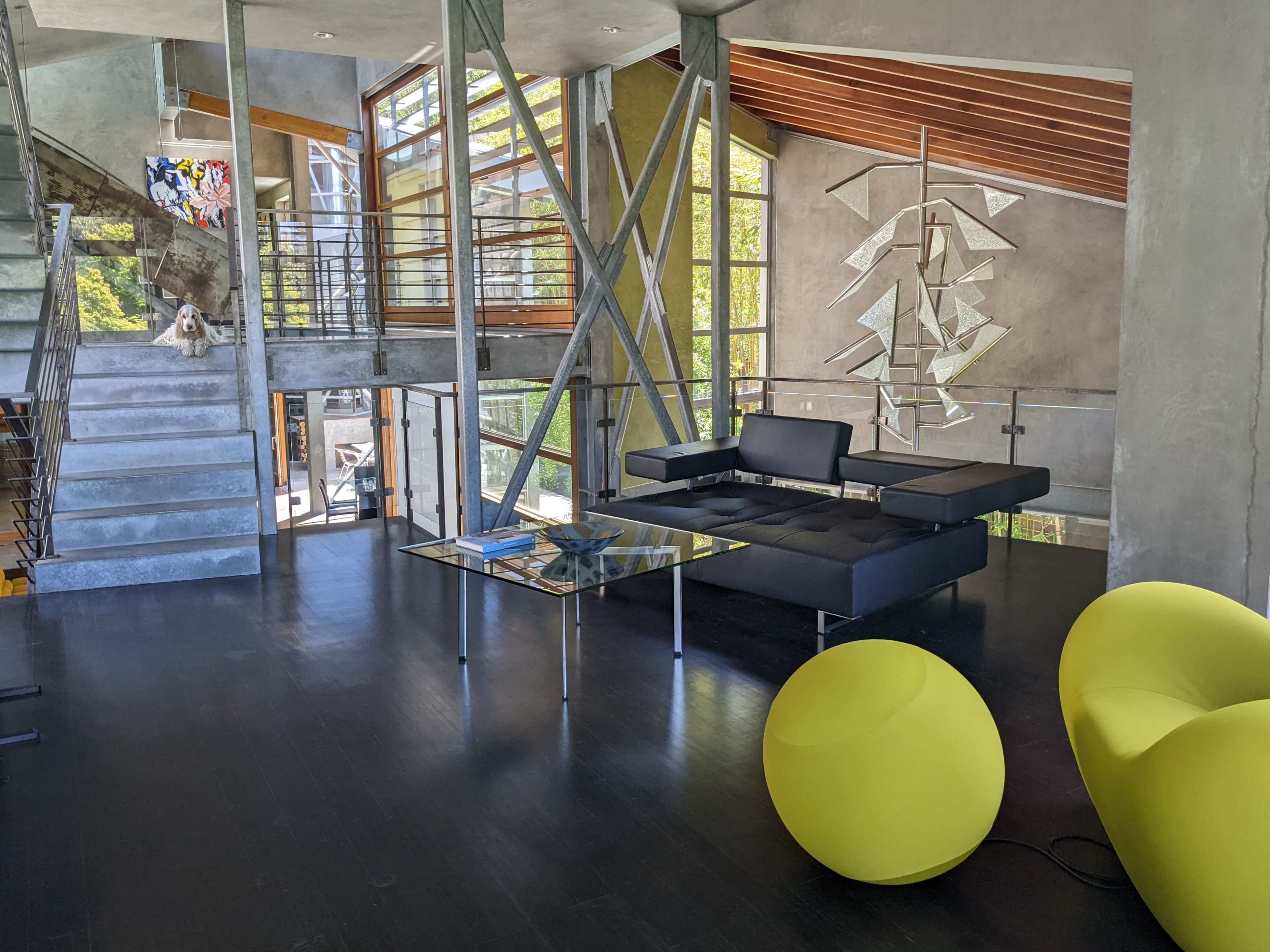 The image shows a modern interior space with large windows, a central glass table, black seating furniture, and a staircase leading to an upper level.