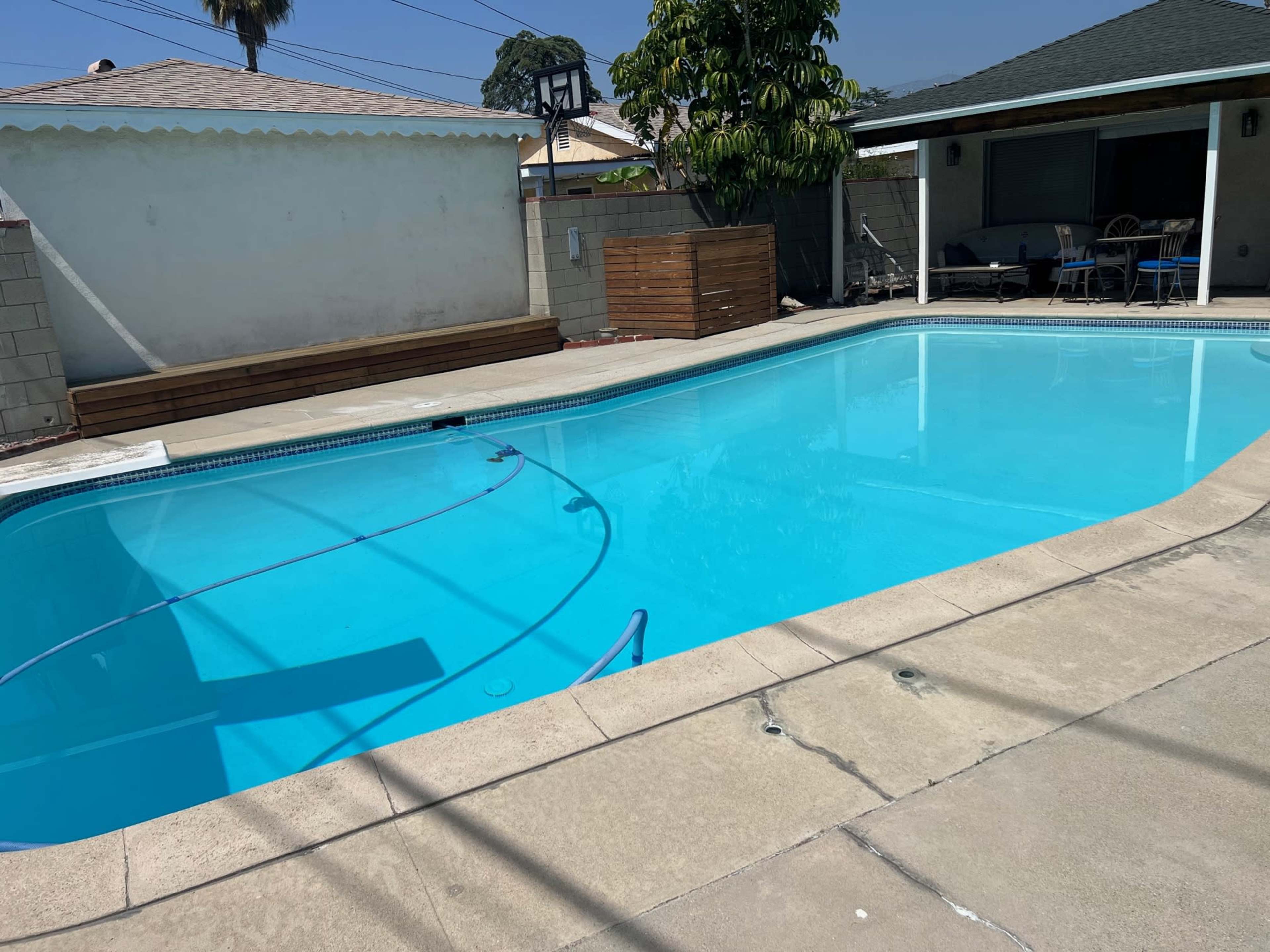 The image shows a rectangular swimming pool with clear blue water, surrounded by concrete pavement, a wooden bench, and a shaded seating area in the background.