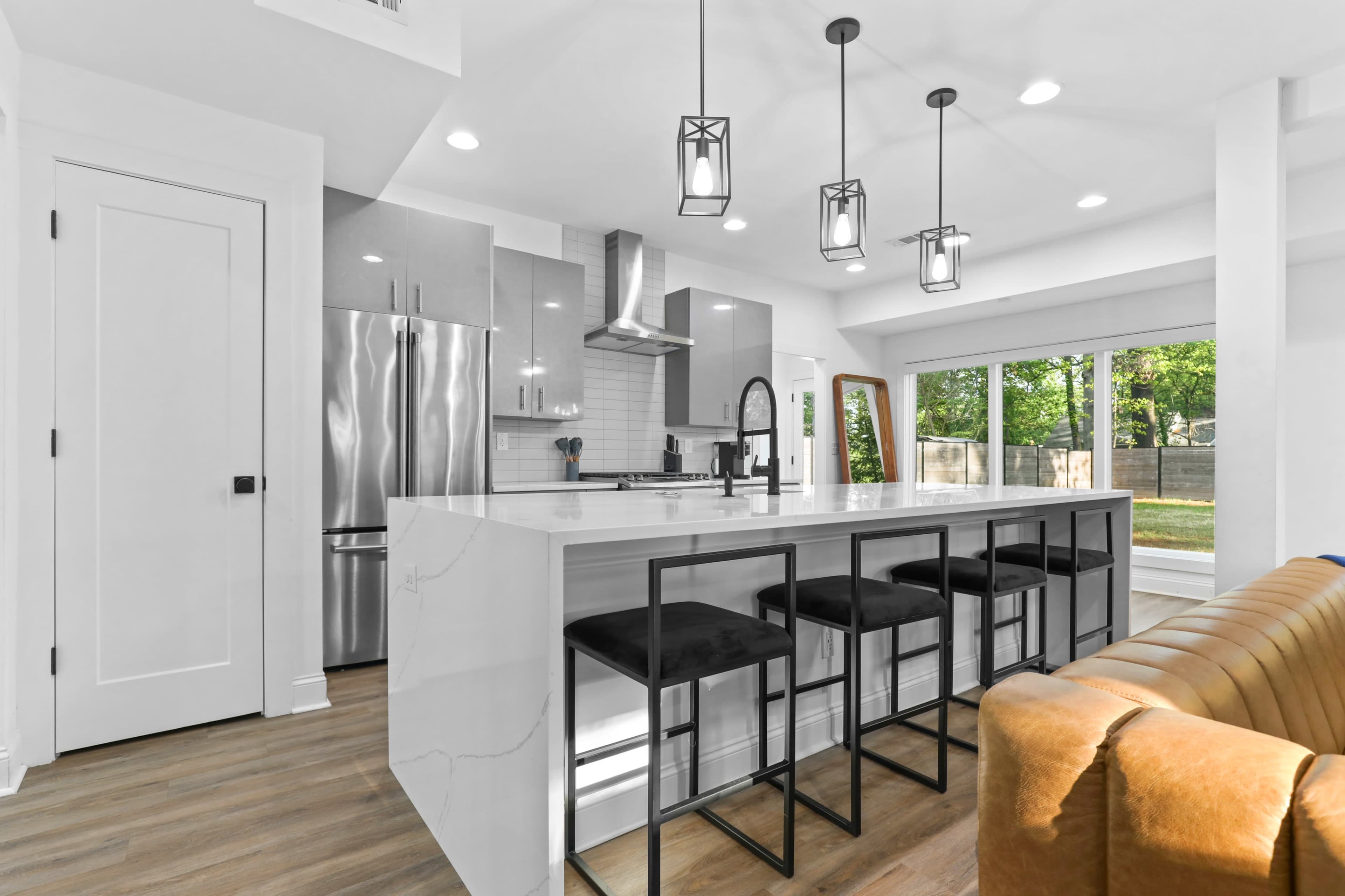A modern kitchen features a large island with seating for four, stainless steel appliances, and a view of the outdoors through large windows.