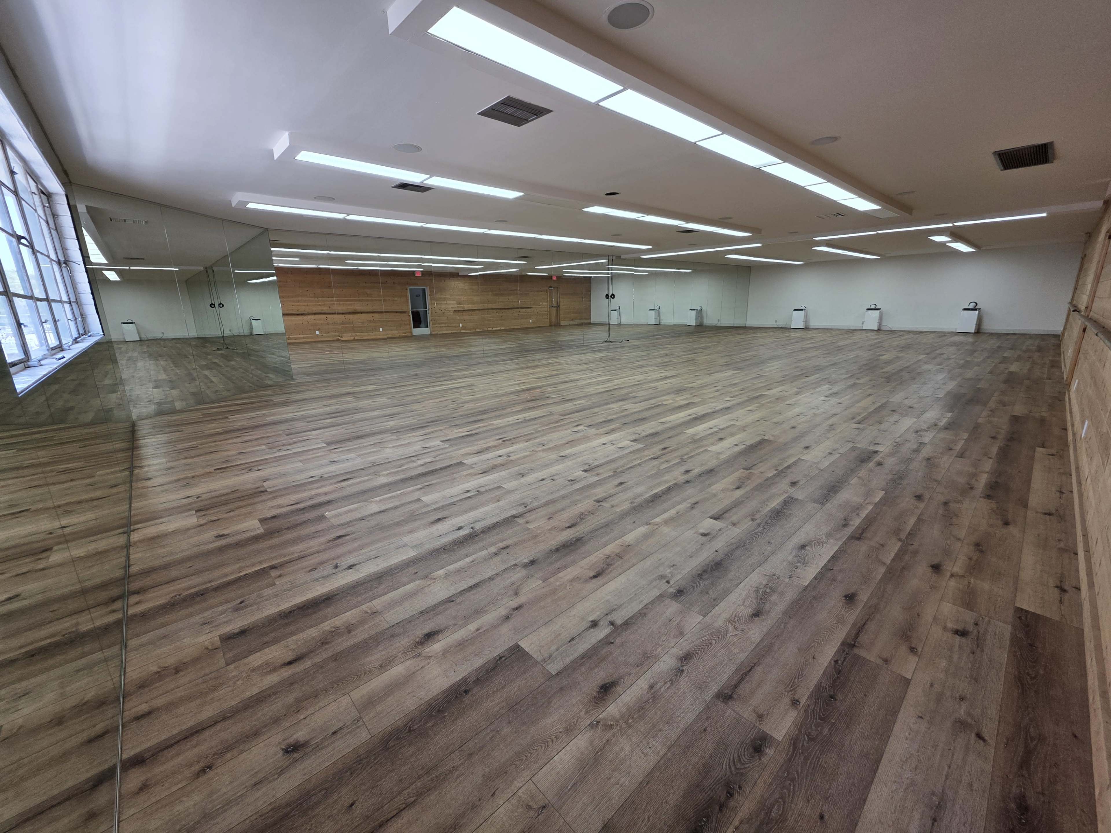 The image shows a large, empty studio space with wooden floors, bright ceiling lights, and mirrors on one wall.