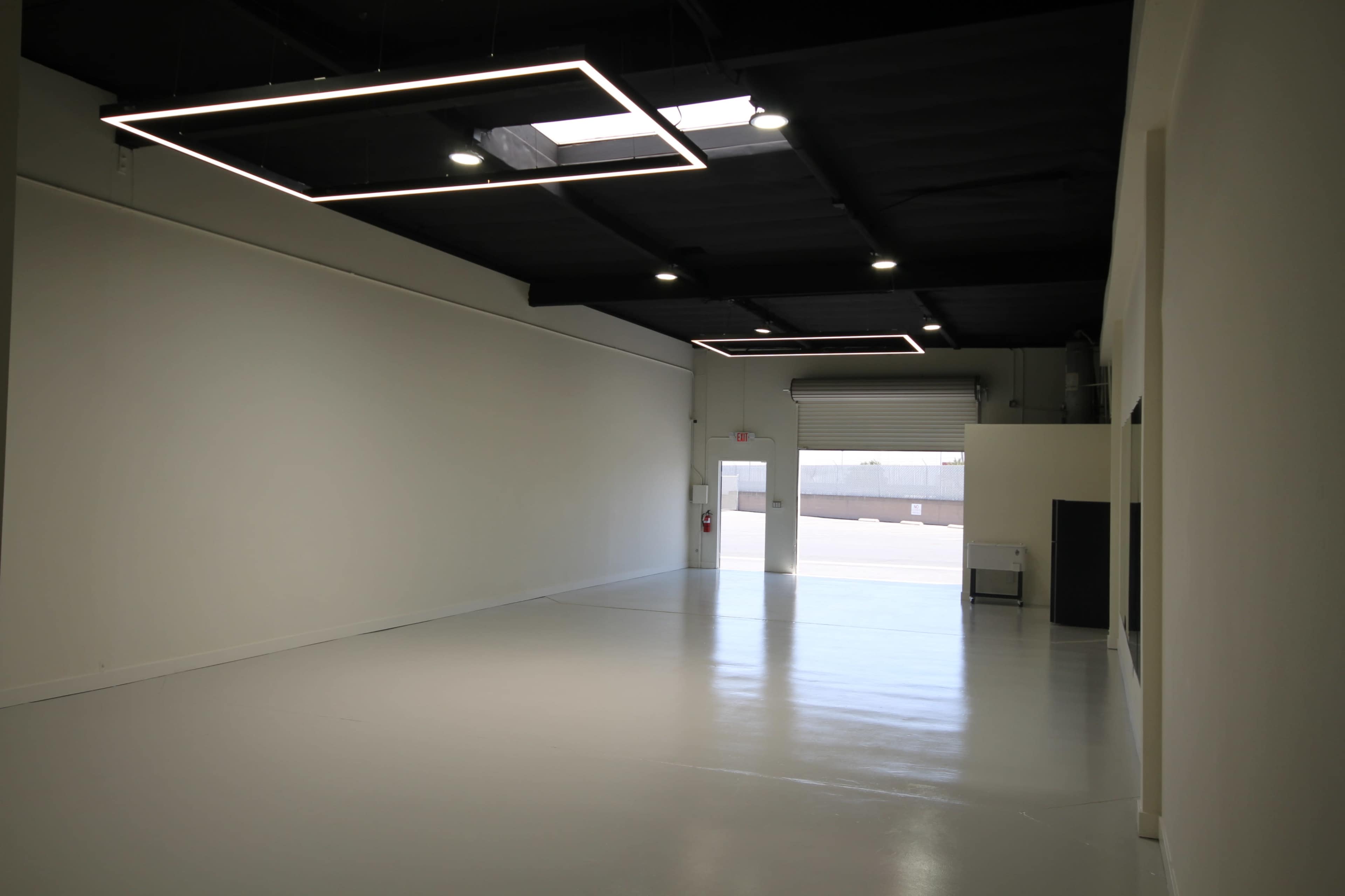 The image shows a spacious, empty room with white walls and a polished floor, featuring modern rectangular light fixtures and a large door opening to the outside.