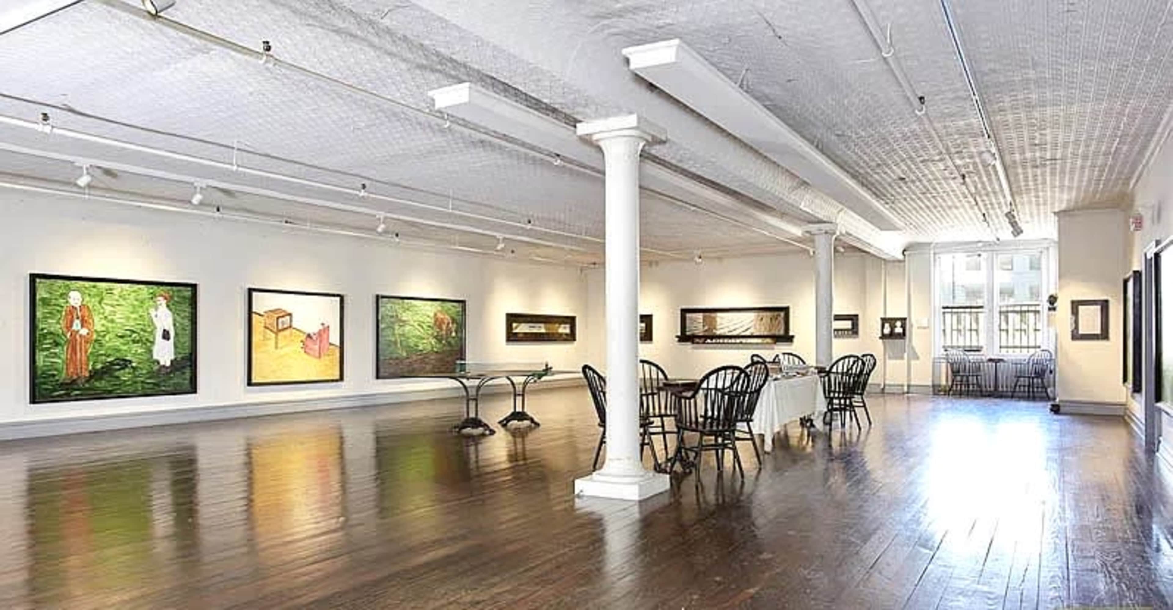 The image shows a spacious art gallery with wooden floors, large paintings on the walls, and a dining table set with chairs in the center.
