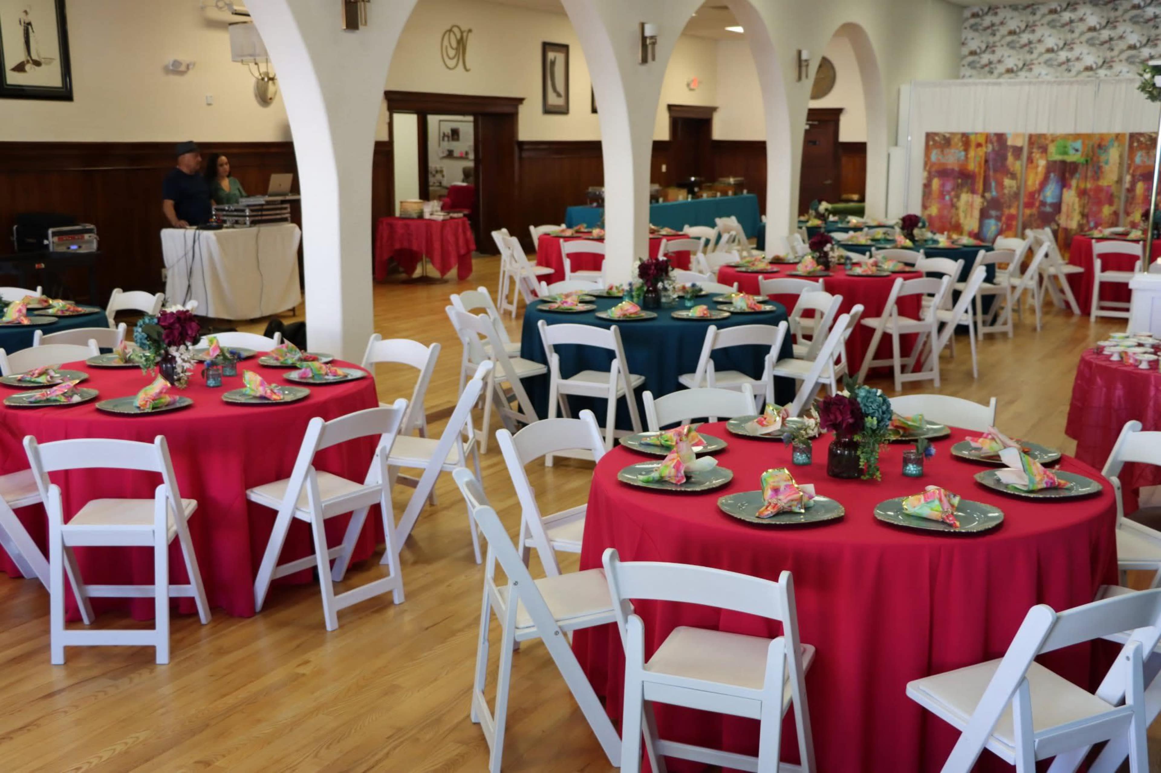 The image shows a spacious event hall set up with round tables covered in red and teal tablecloths, each arranged with plates and floral centerpieces.