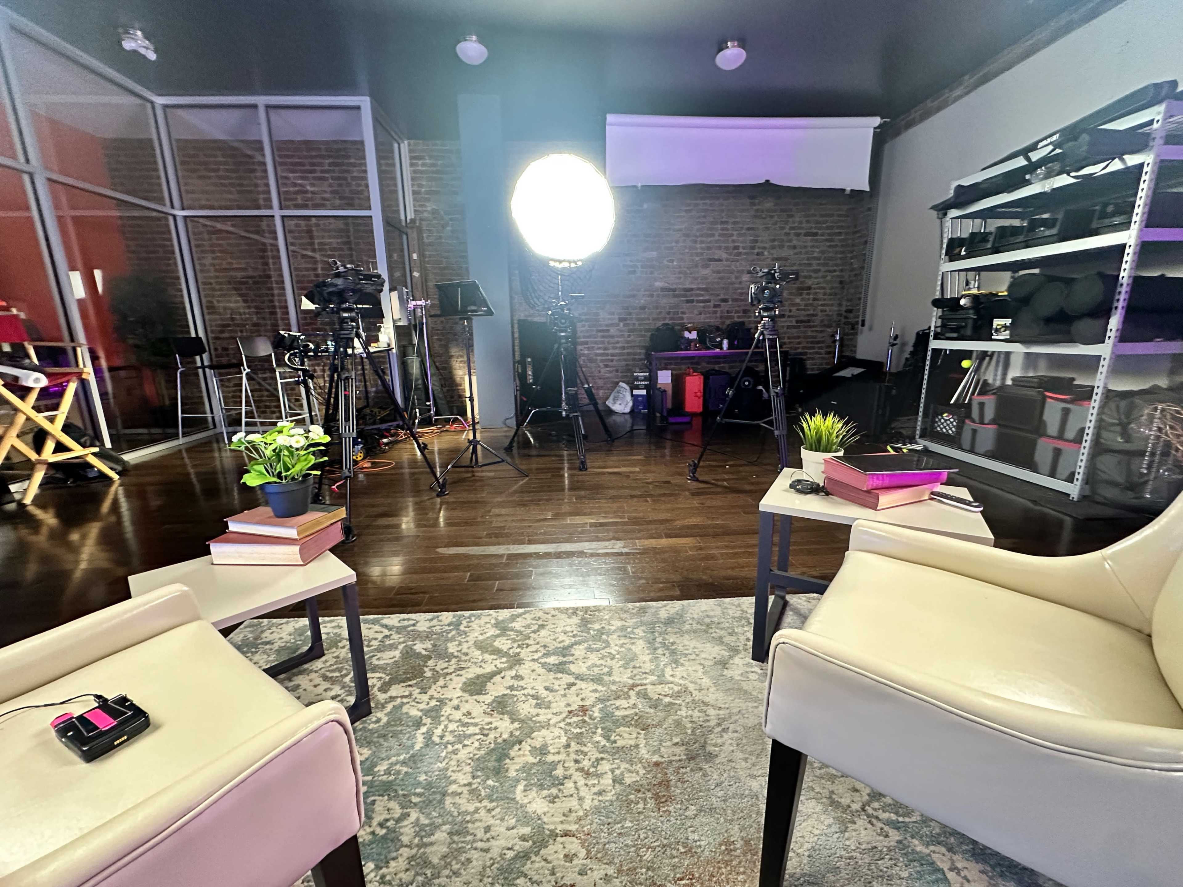 A well-lit studio setup with two chairs, a rug, and various filming equipment in the background.