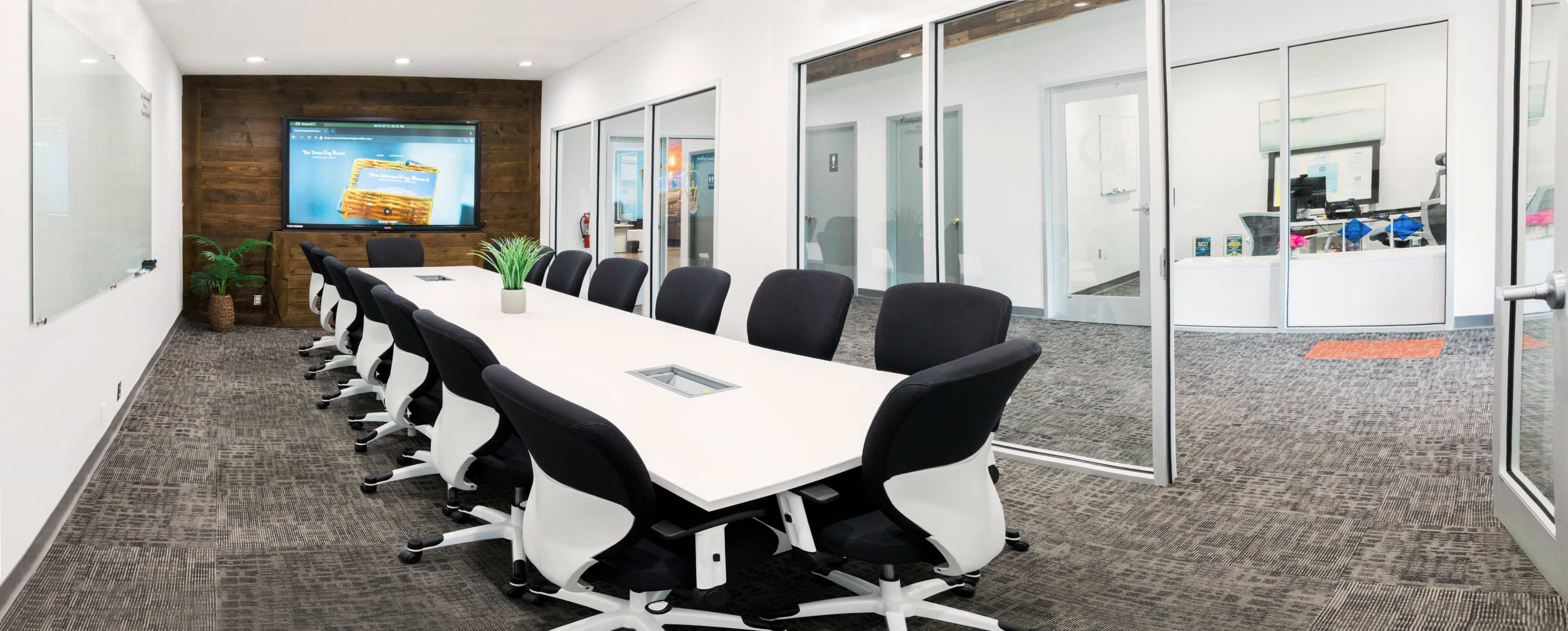 A modern conference room features a long white table surrounded by black ergonomic chairs, with glass walls and a digital screen displaying content.