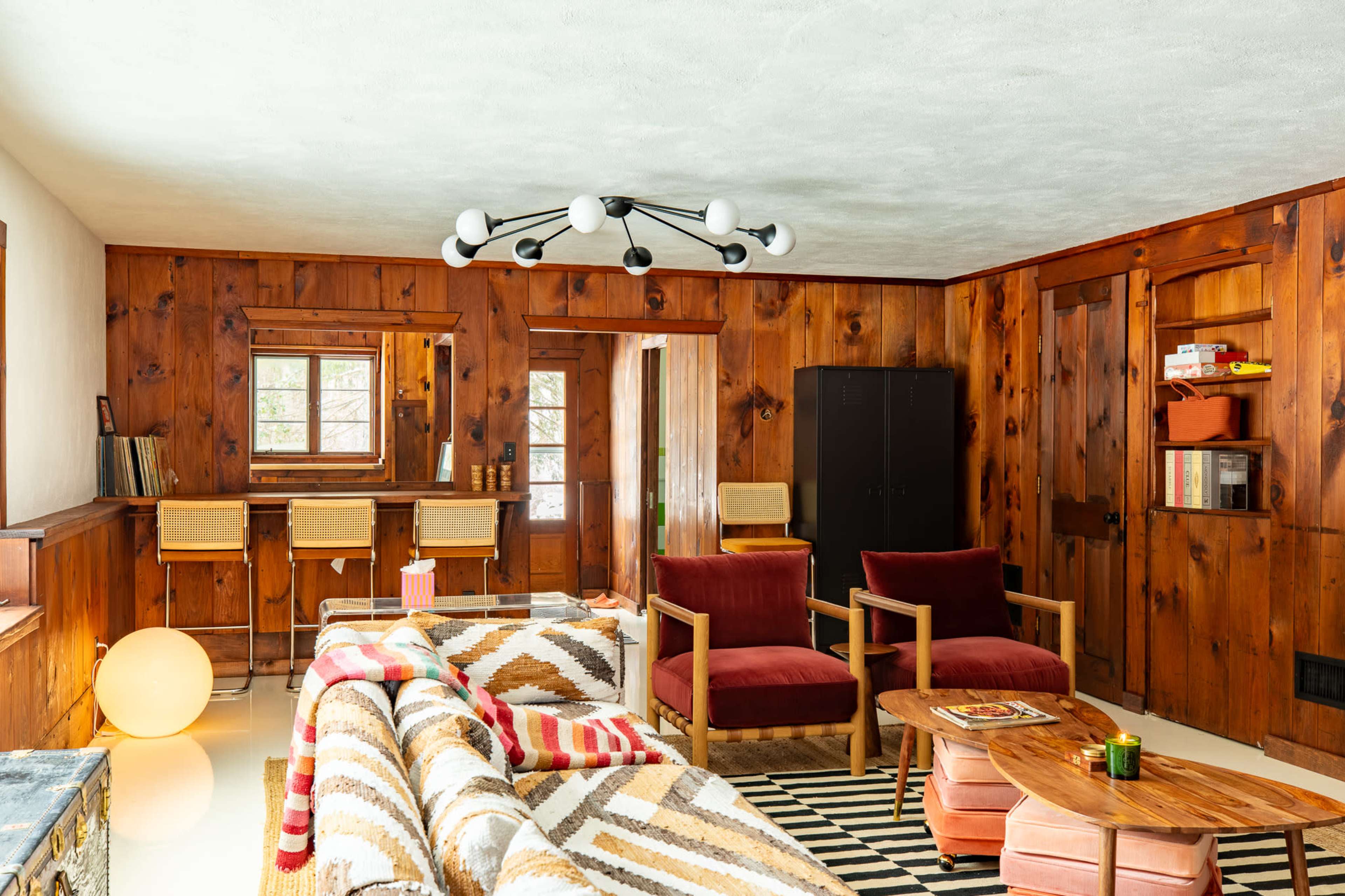 The image shows a cozy, wood-paneled living room featuring striped seating, a circular coffee table, and a unique light fixture.
