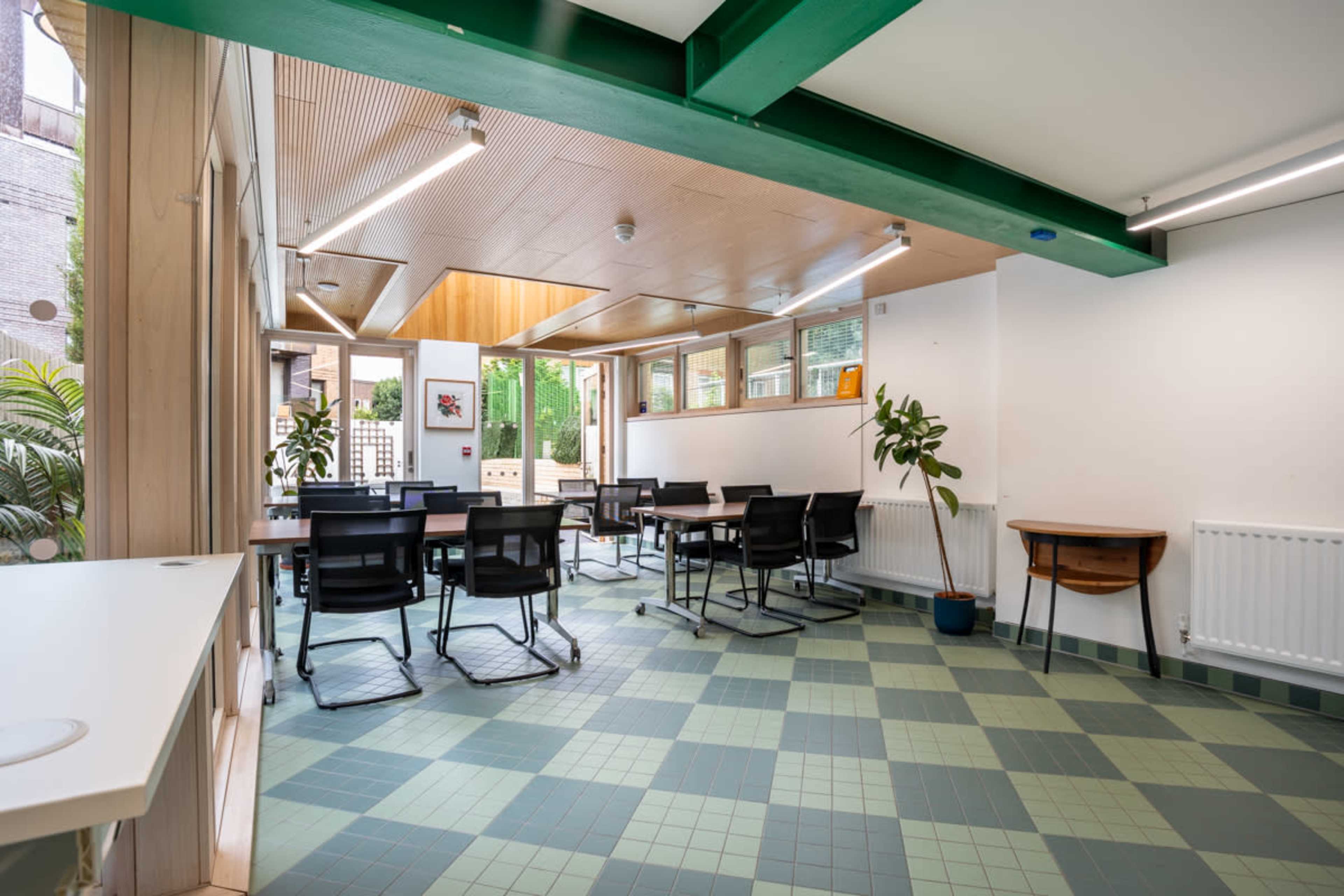 A well-lit multi-purpose room with tables and chairs arranged for meetings, featuring large windows overlooking a garden.