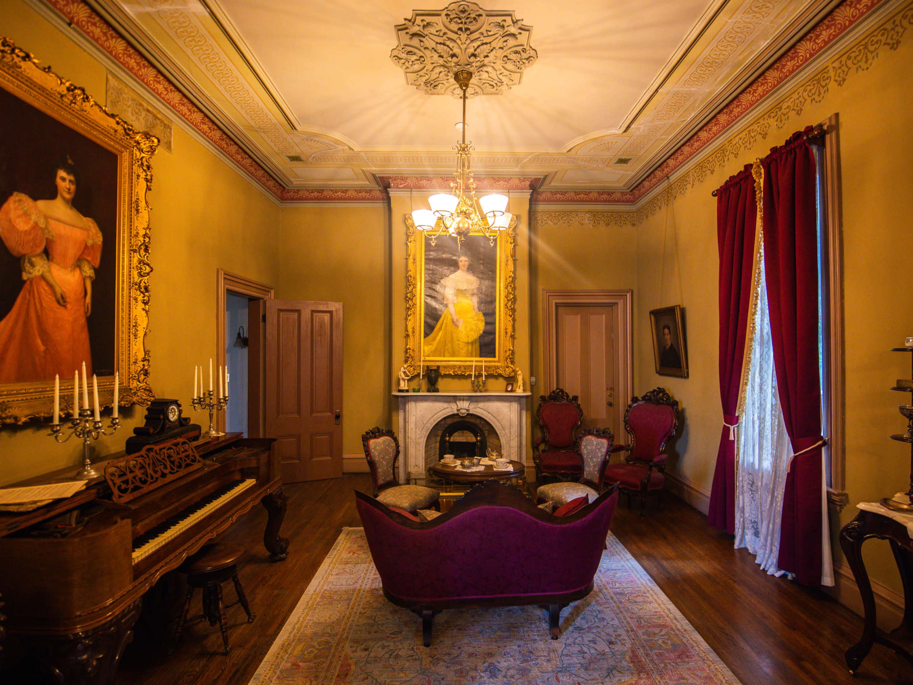A vintage sitting room featuring a grand piano, a purple upholstered sofa, and elegant portraits on the walls.
