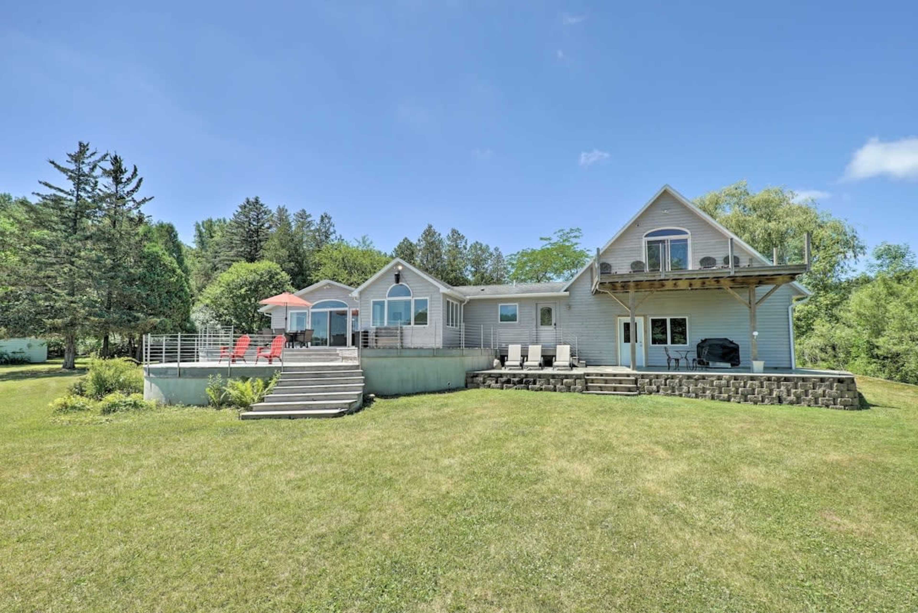 A large, two-story house with a deck and patio furniture is situated on a grassy lawn surrounded by trees.