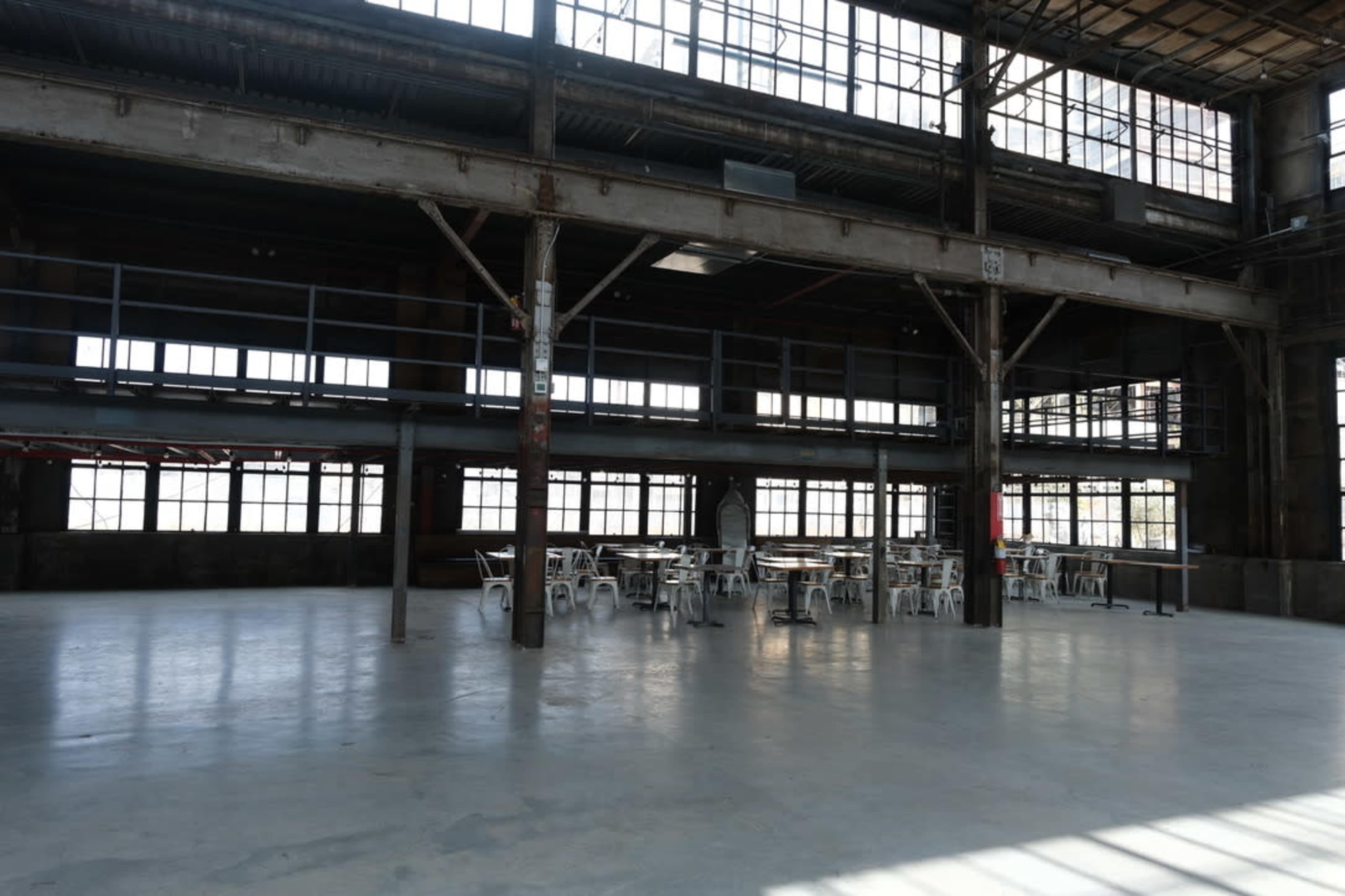 The image shows a spacious, industrial-style interior with large windows, exposed beams, and several tables and chairs set up in the area.