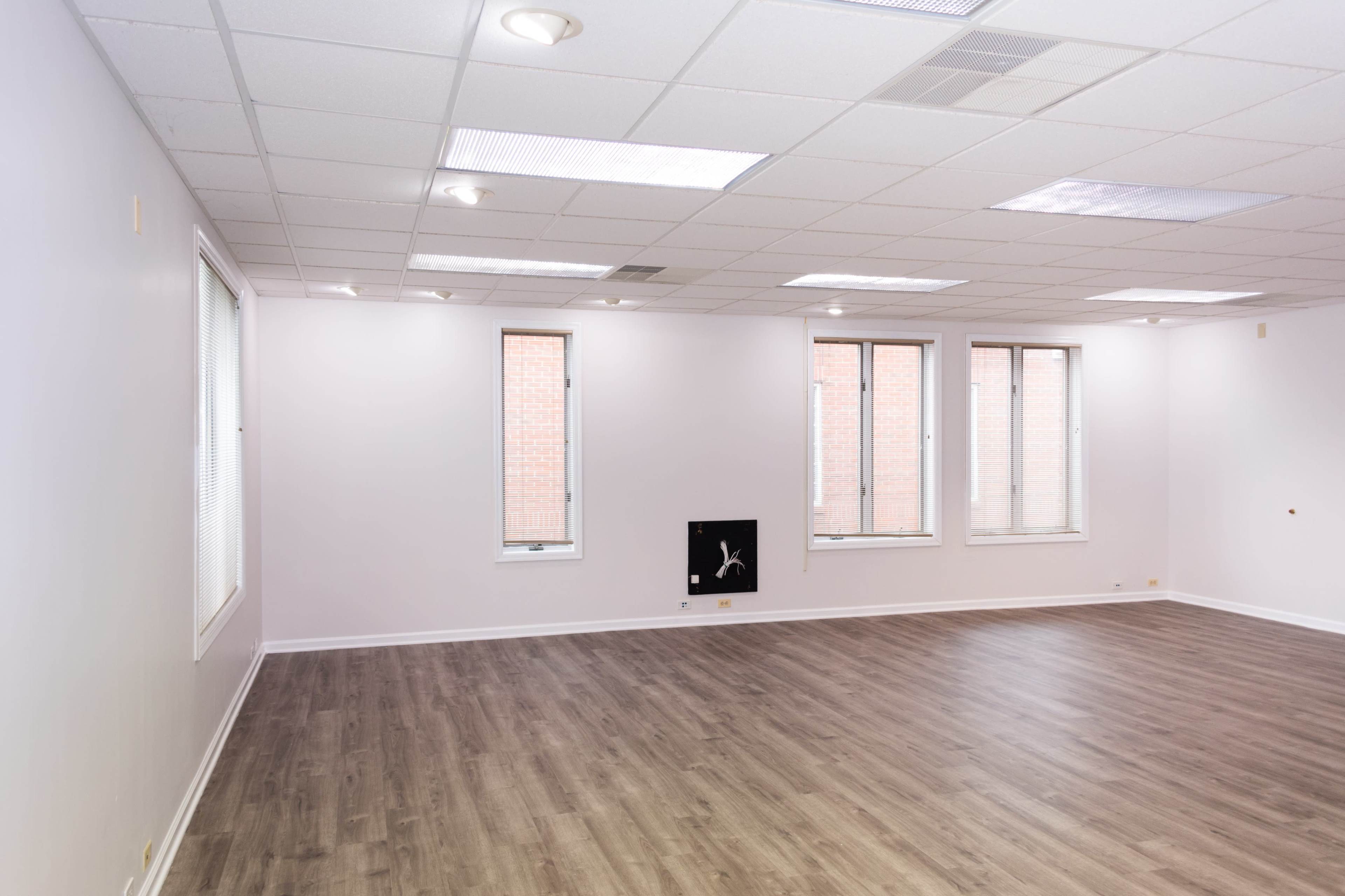 An empty room with light-colored walls and hardwood flooring features several windows along one side.