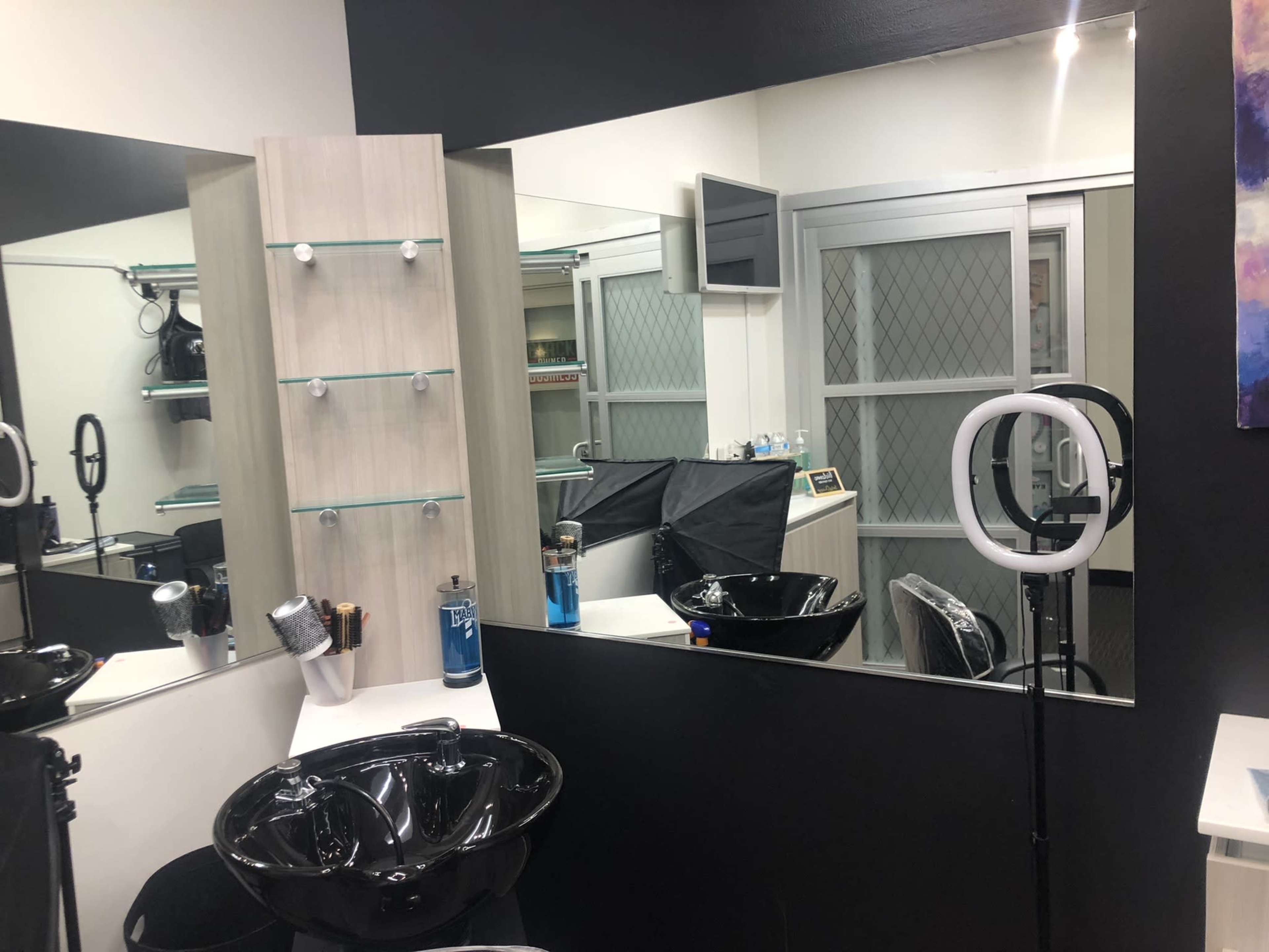A hair salon interior featuring a black sink, mirrors, and a ring light setup in a well-lit space.