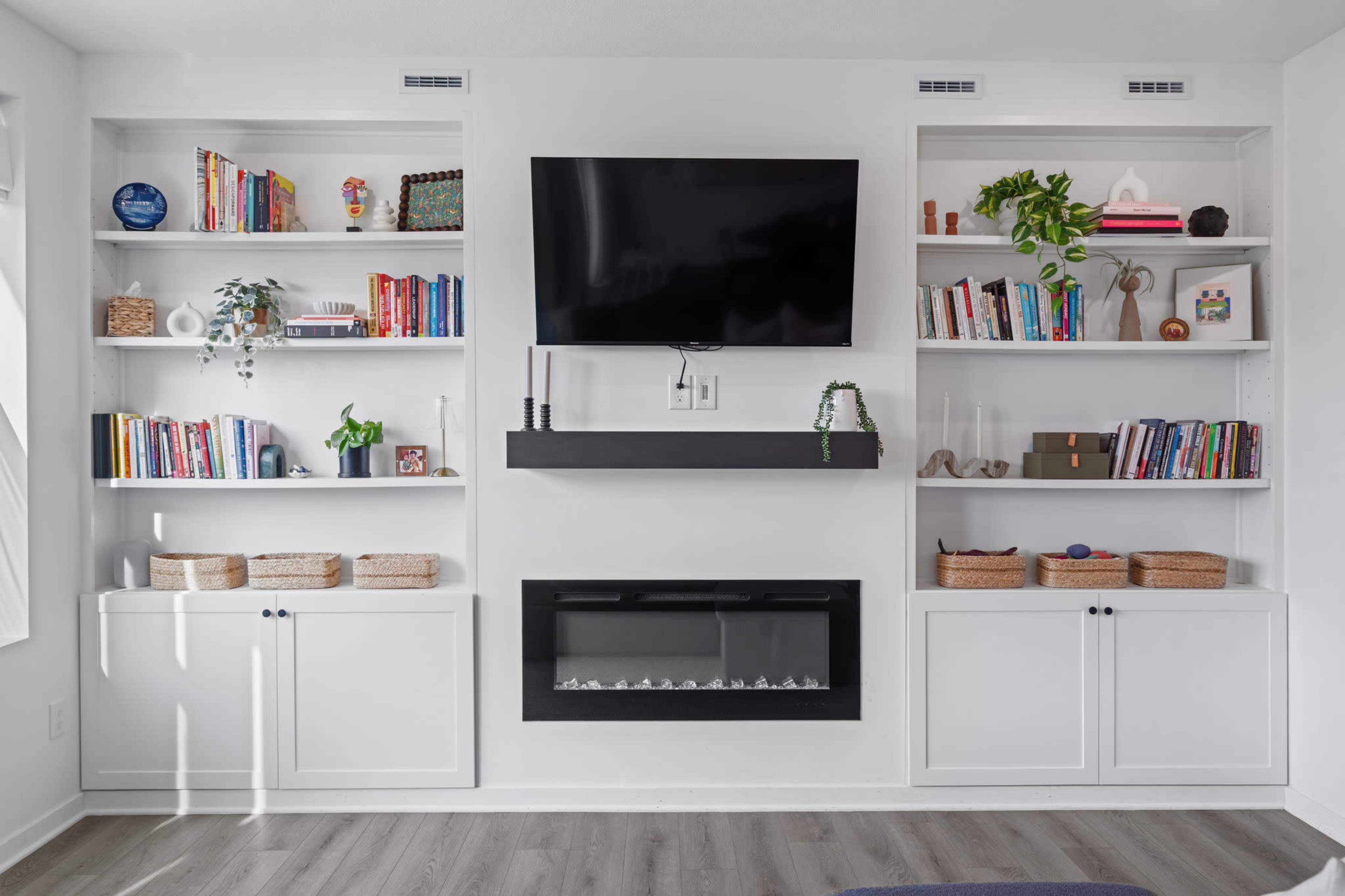 The image shows a living room wall featuring built-in shelving with books, decorative items, and a mounted television above a modern electric fireplace.