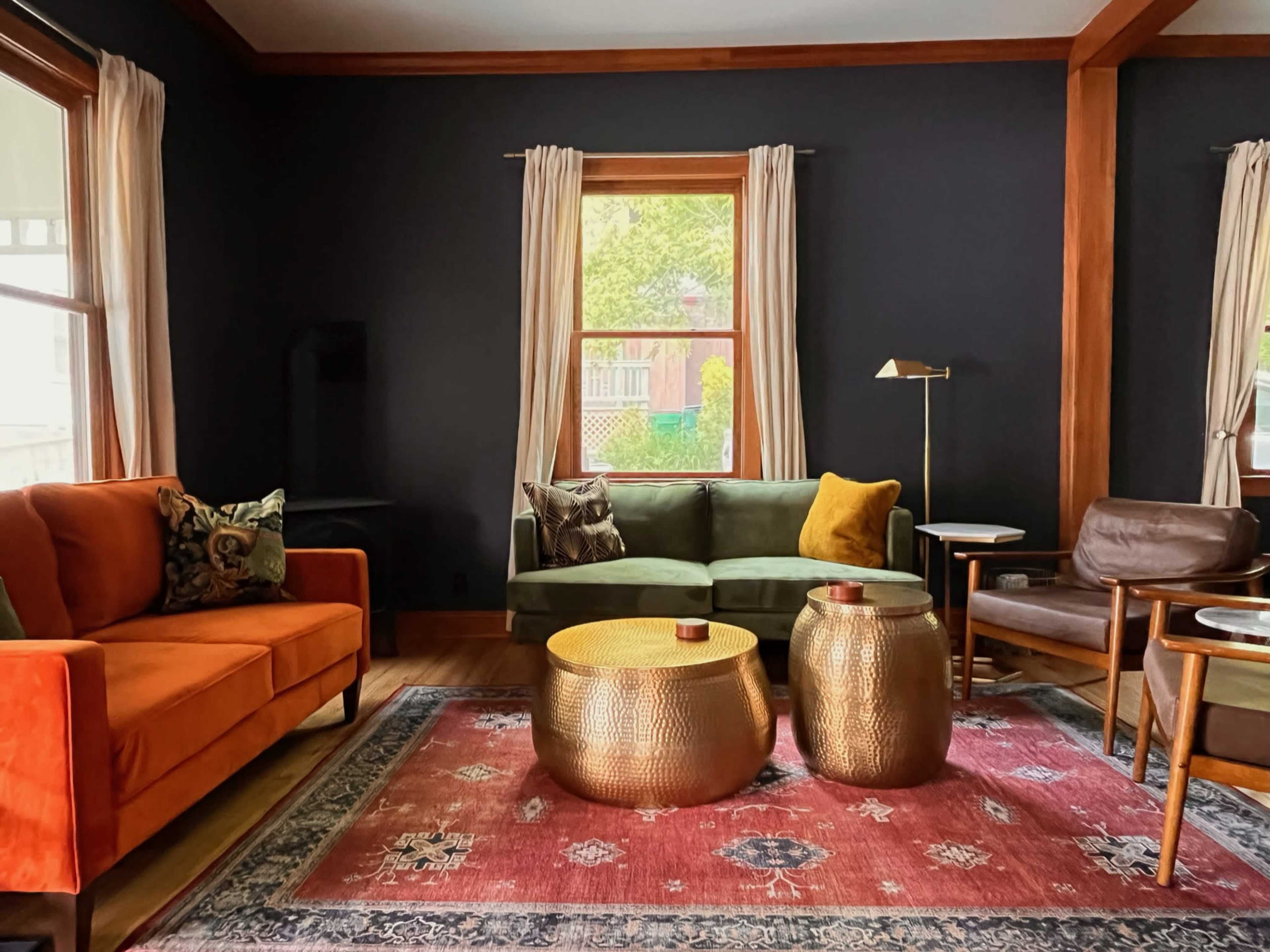 A living room features an orange sofa, a green couch, two gold accent tables, and a patterned rug under a window with sheer curtains.