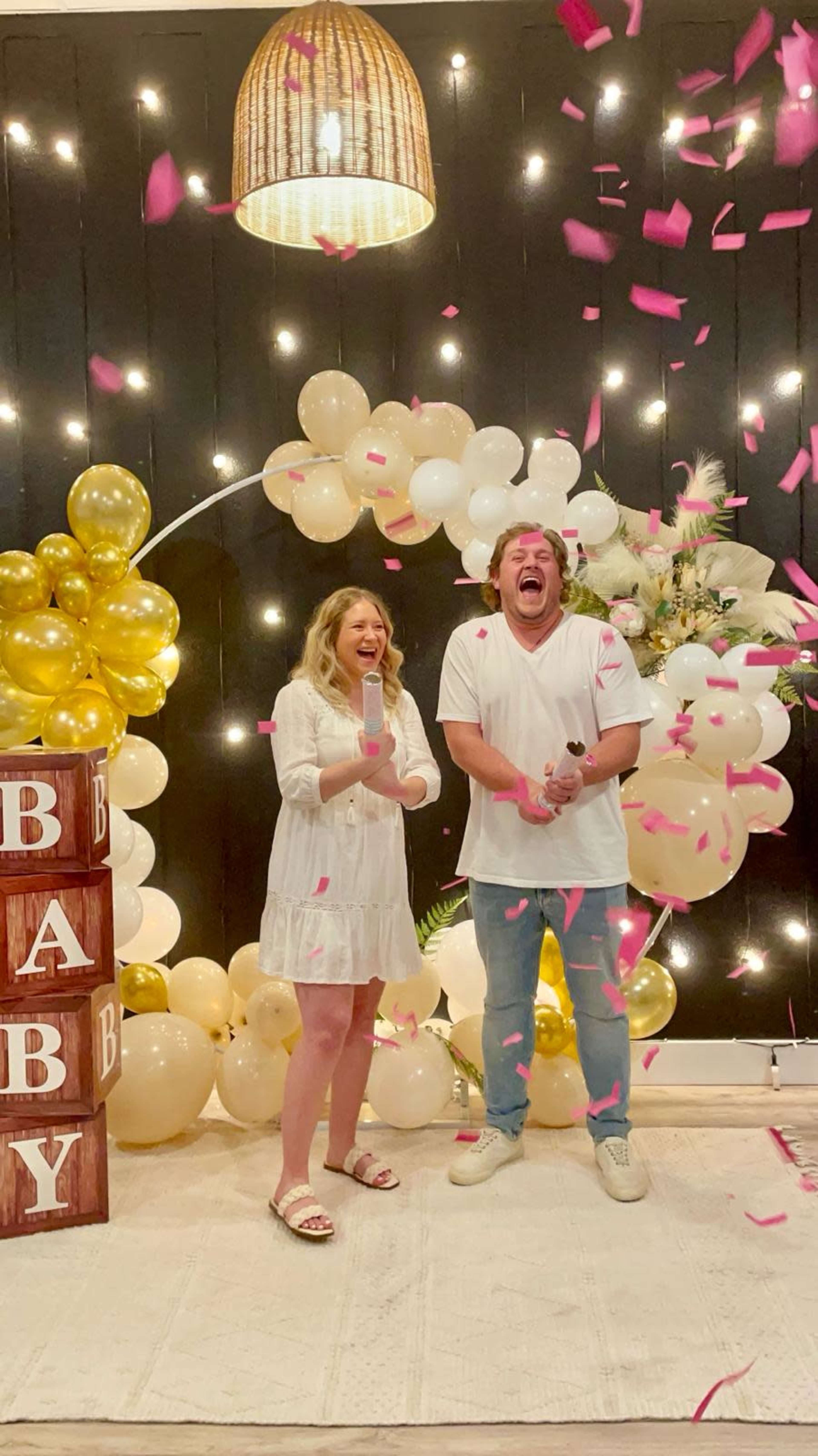 A couple stands joyfully among balloons and decorations while confetti falls around them during a celebration.
