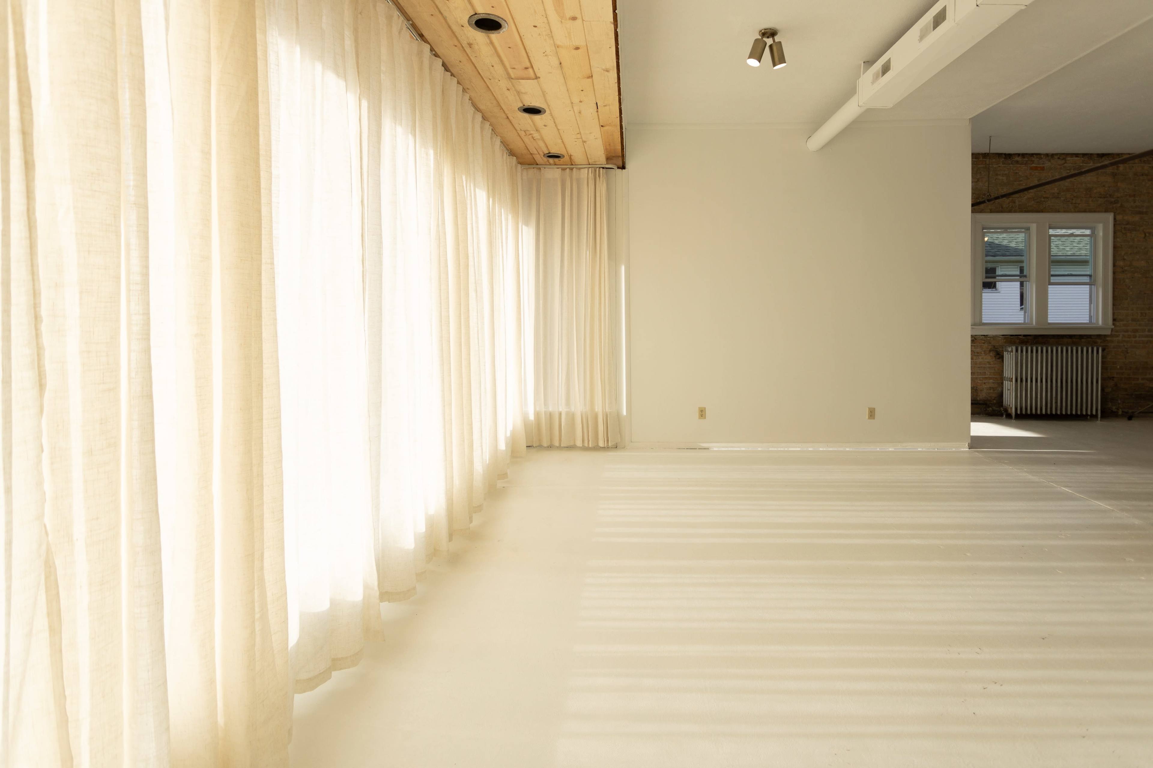 A spacious room with light-colored walls features large windows covered by sheer curtains, allowing sunlight to cast soft shadows on the floor.