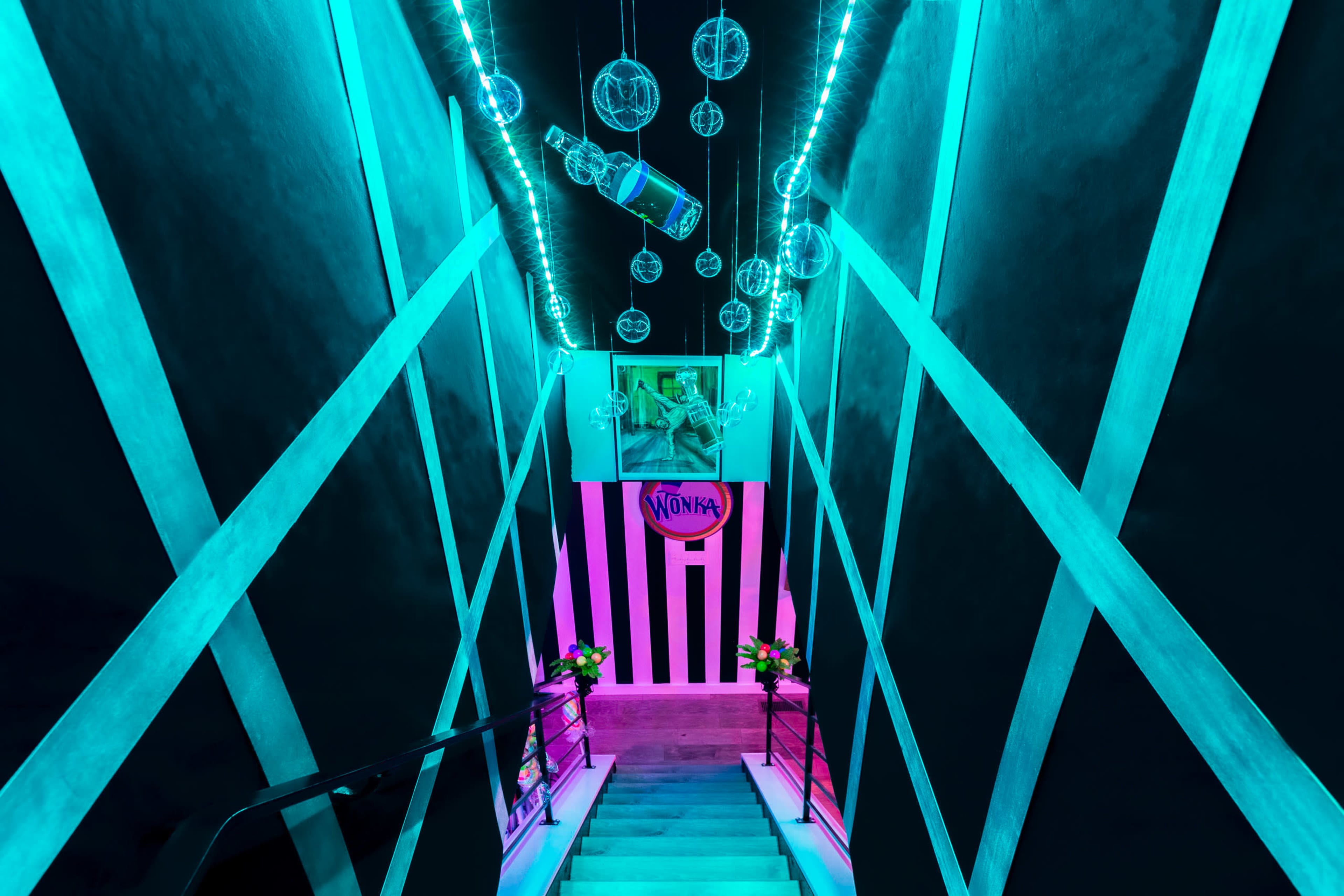 A staircase illuminated by blue and pink LED lights, with hanging glass orbs above and a "Wonka" sign on a wall.