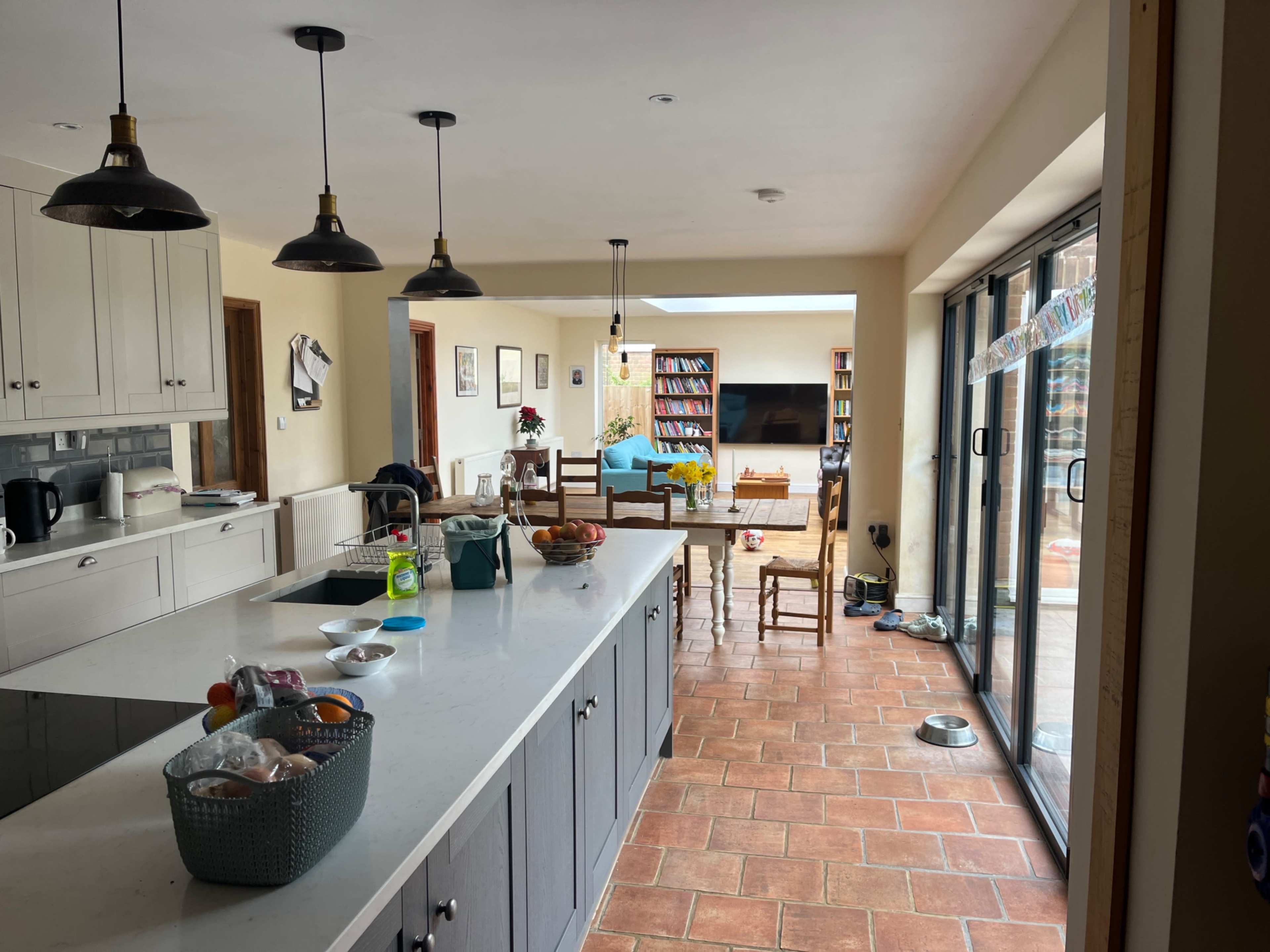 The image shows a modern kitchen and dining area featuring a large island, wooden dining table, brown tiled floor, and glass doors leading to an outdoor space.