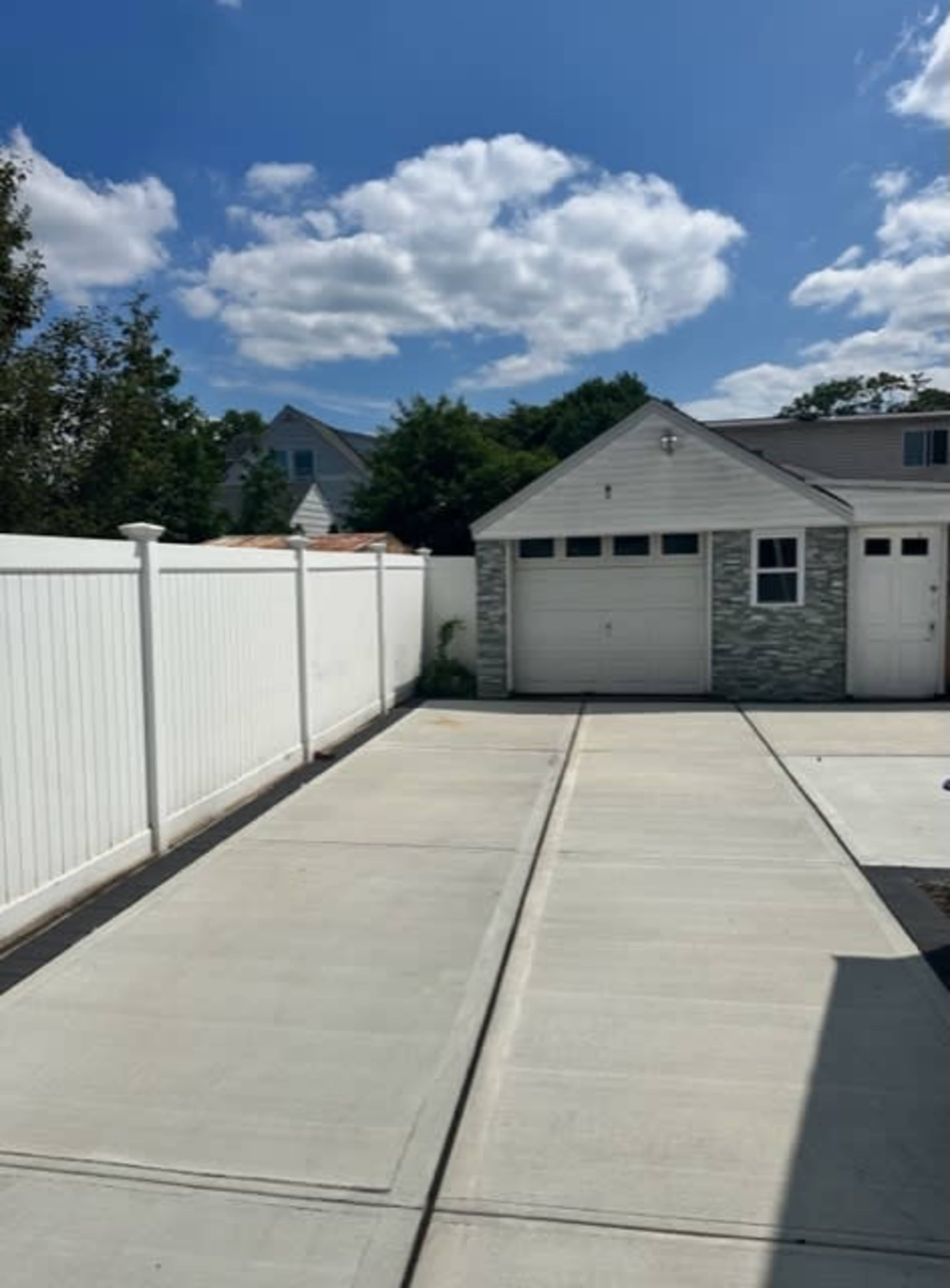 A concrete driveway leading to a garage, flanked by a tall white fence and under a partly cloudy sky.
