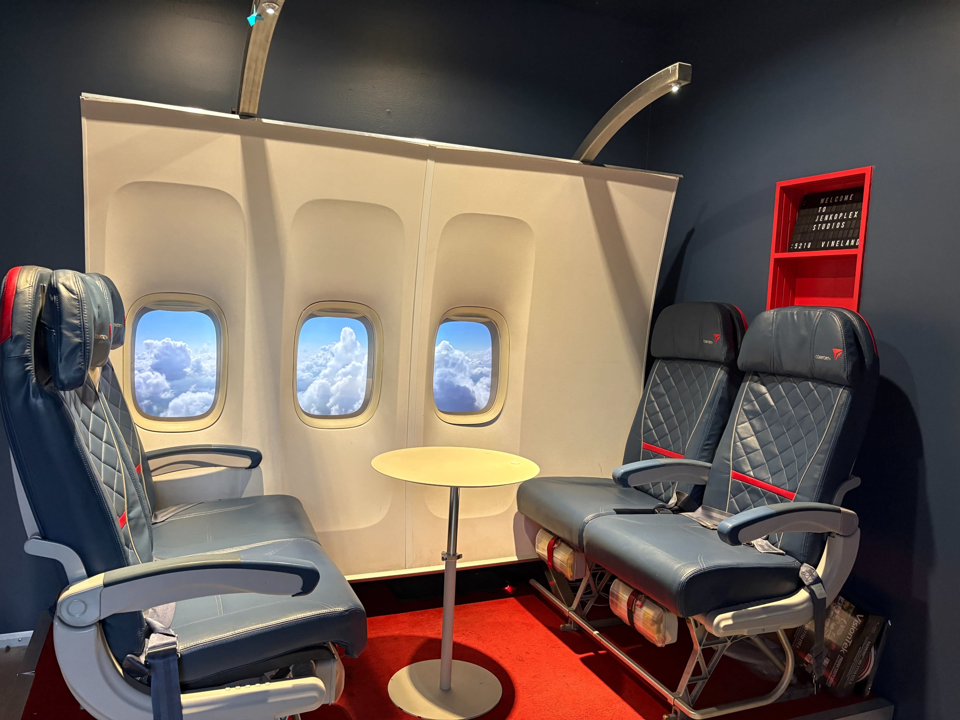 The image shows a mock-up of an airplane interior featuring two airplane seats with a small table between them and a backdrop of windows displaying clouds.