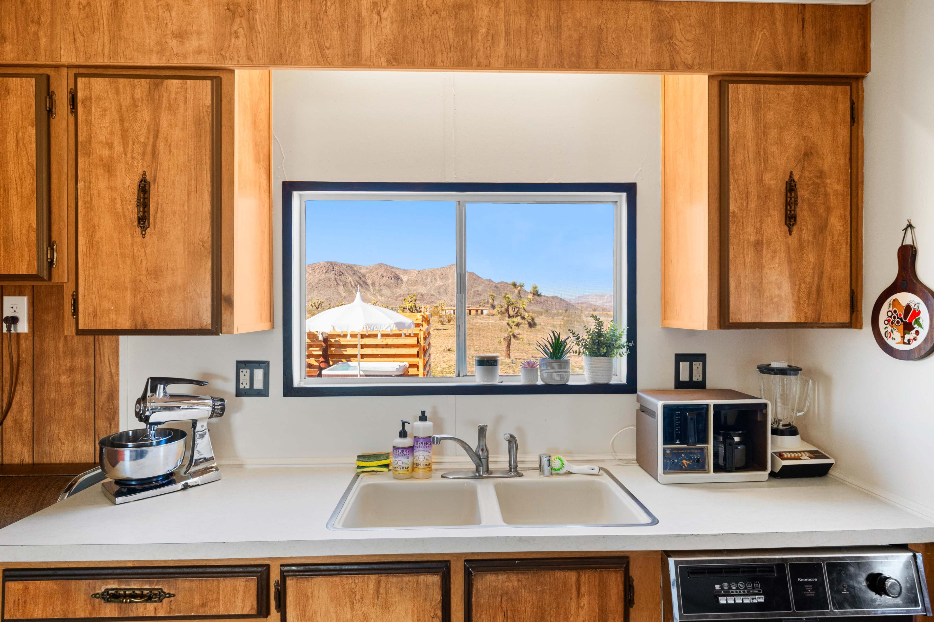 A kitchen with wooden cabinets and a clean countertop features a window displaying a desert landscape and outdoor seating.