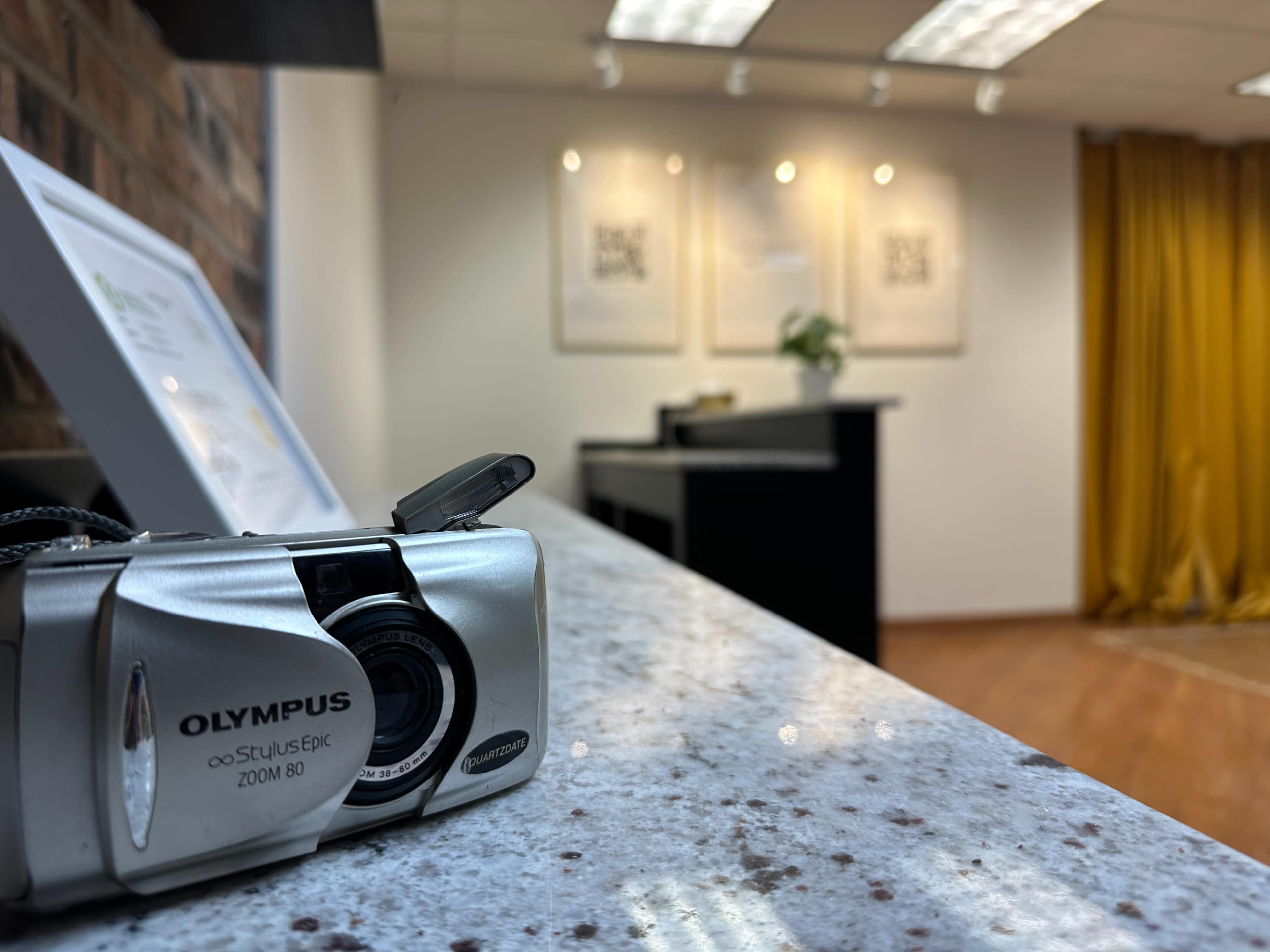 A silver Olympus camera sits on a granite counter in a brightly lit room with yellow curtains and framed artworks on the wall.
