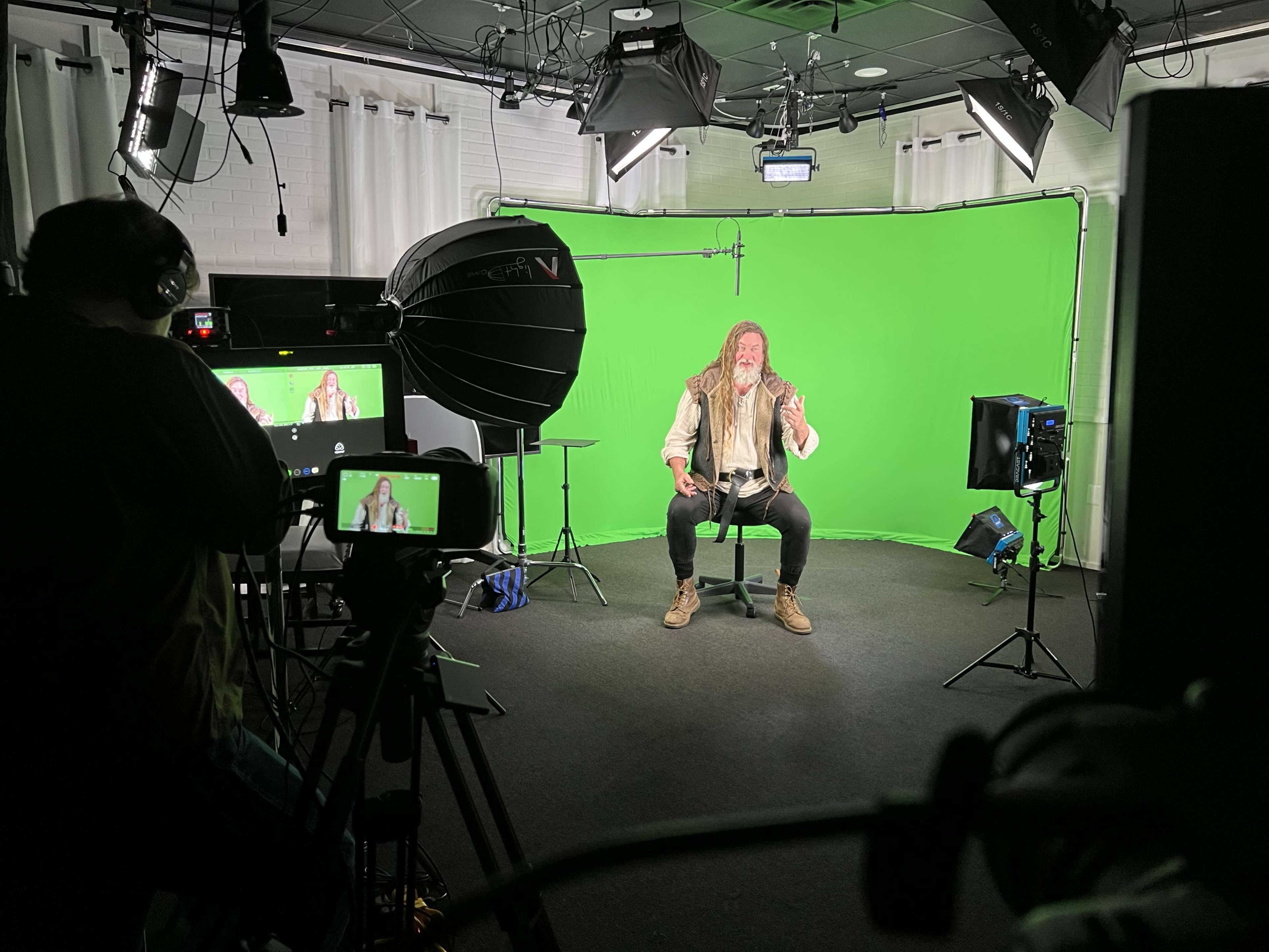 A man with long hair and a beard sits on a stool in front of a green screen while being filmed by multiple cameras in a studio.
