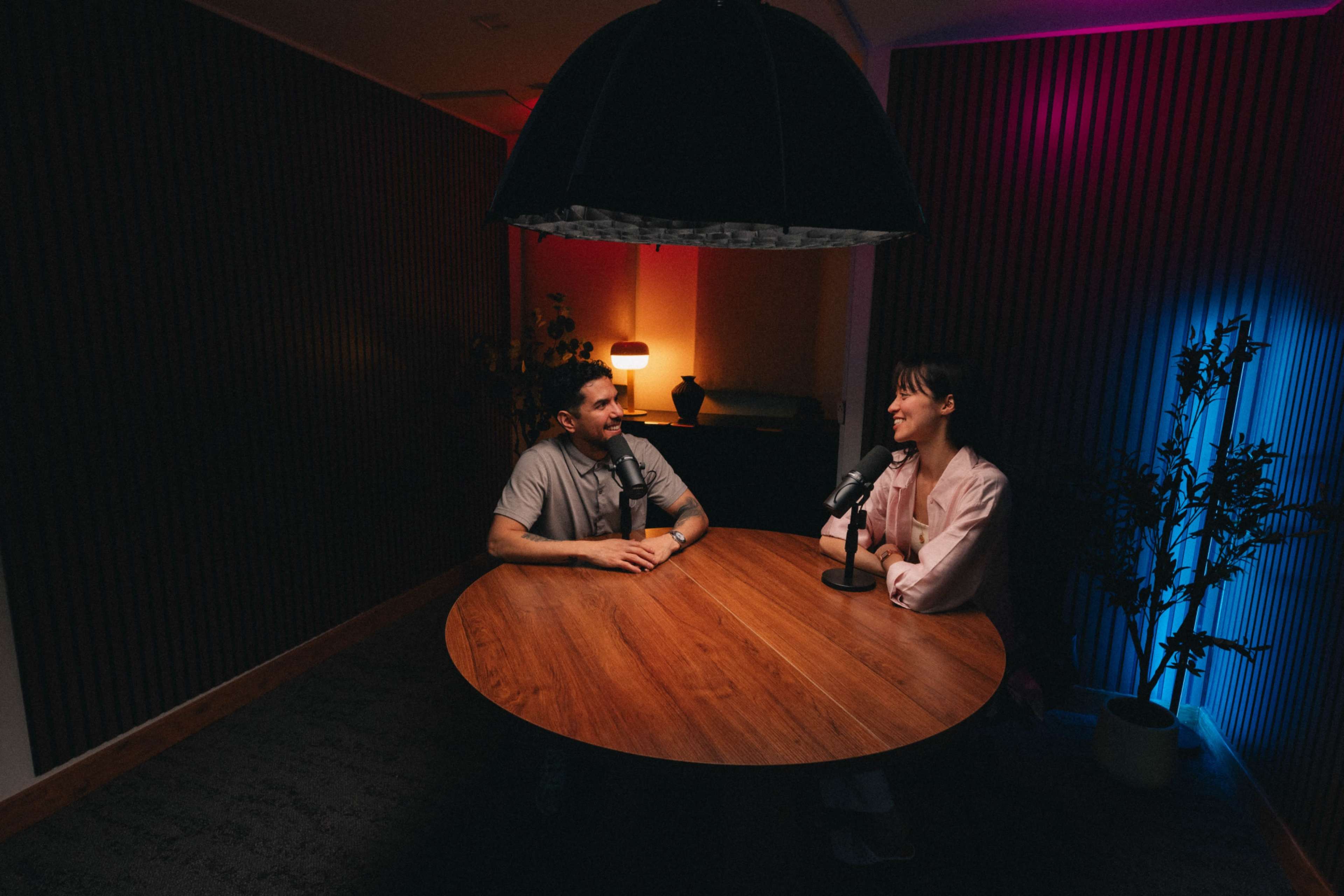 Two people are seated at a round wooden table in a softly lit room, with microphones in front of them and colorful lighting along the walls.